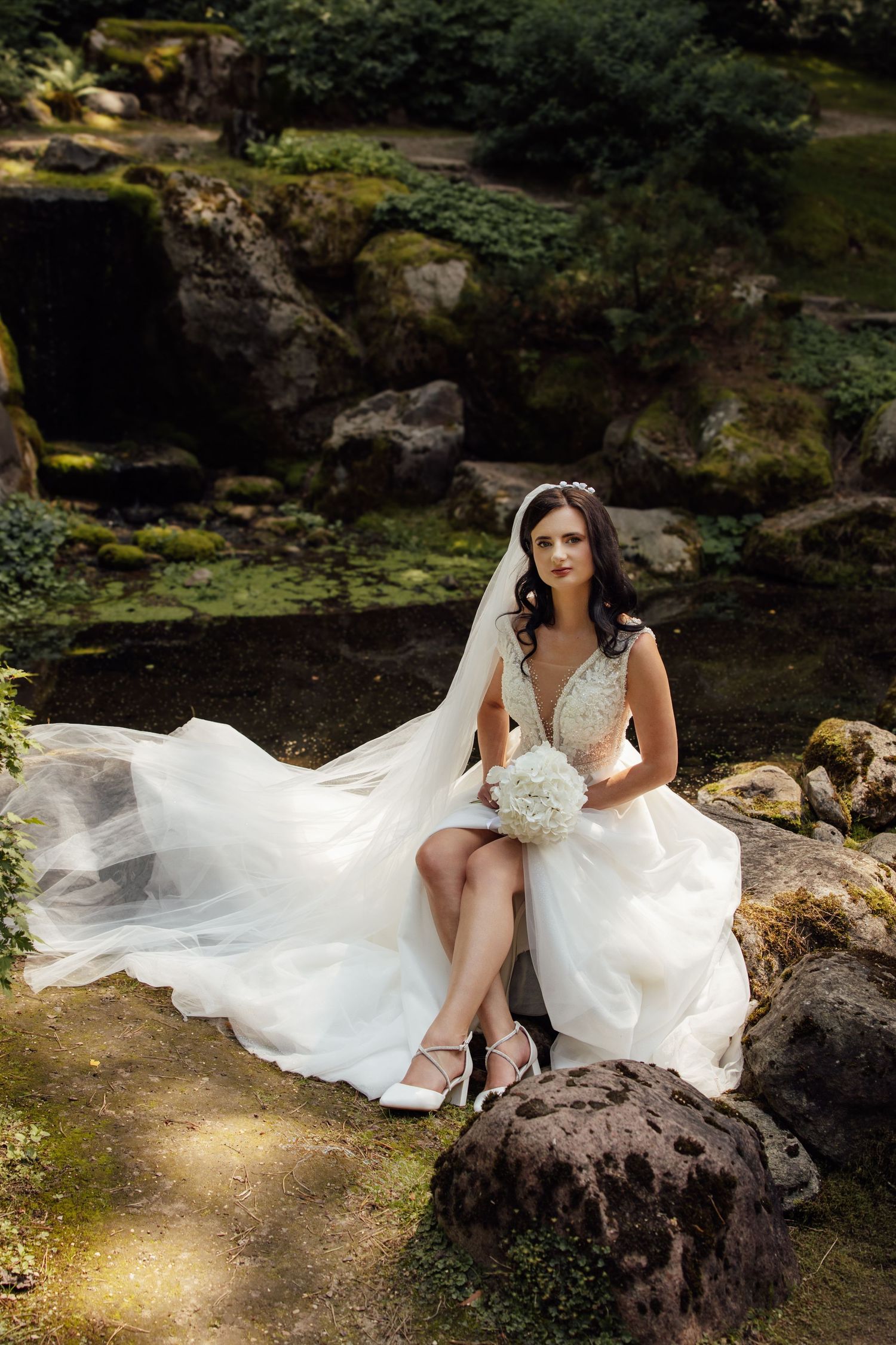 Bride in white lace gown with flowing veil sits on rocks in lush garden holding white bouquet.