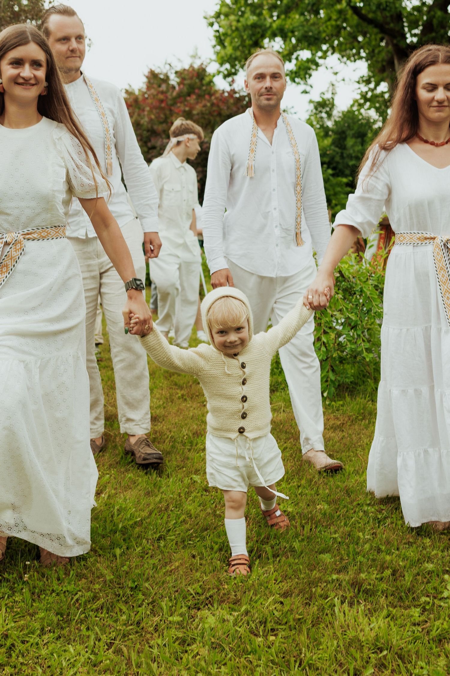 Group of adults in white traditional folk clothing walk outdoors holding hands with a smiling toddler.