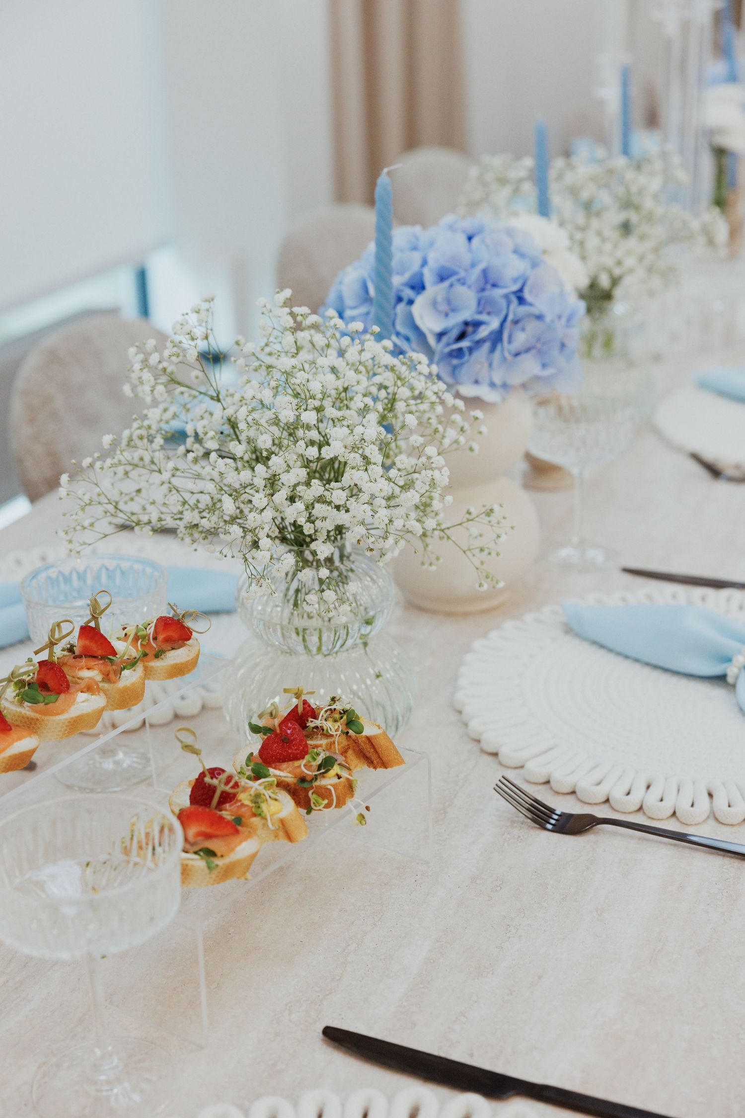 Elegant table setting with baby's breath flowers, blue hydrangeas, and appetizers on white tablecloth.