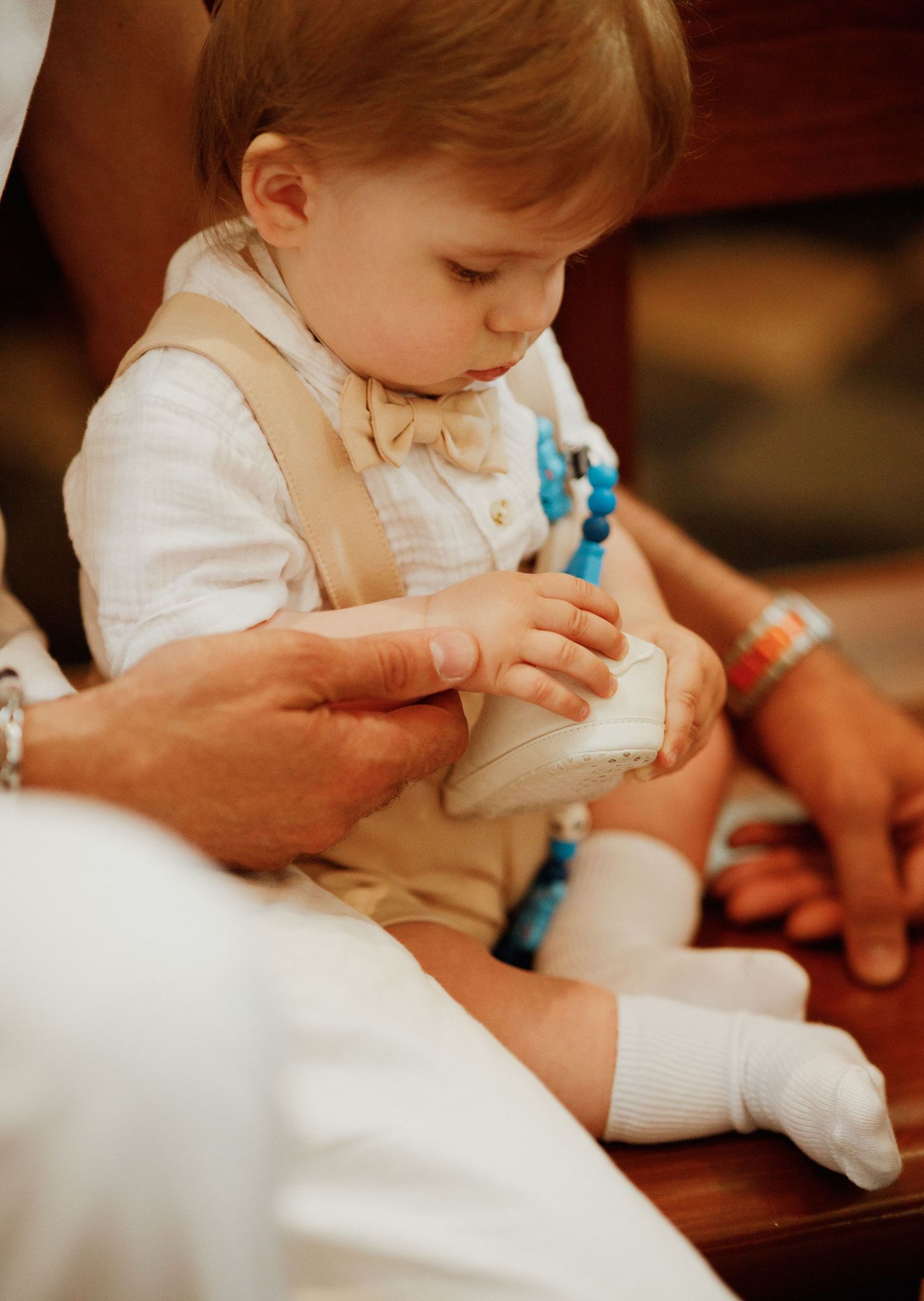 A small child in white and beige formal attire sits while someone helps put on their socks and shoes.