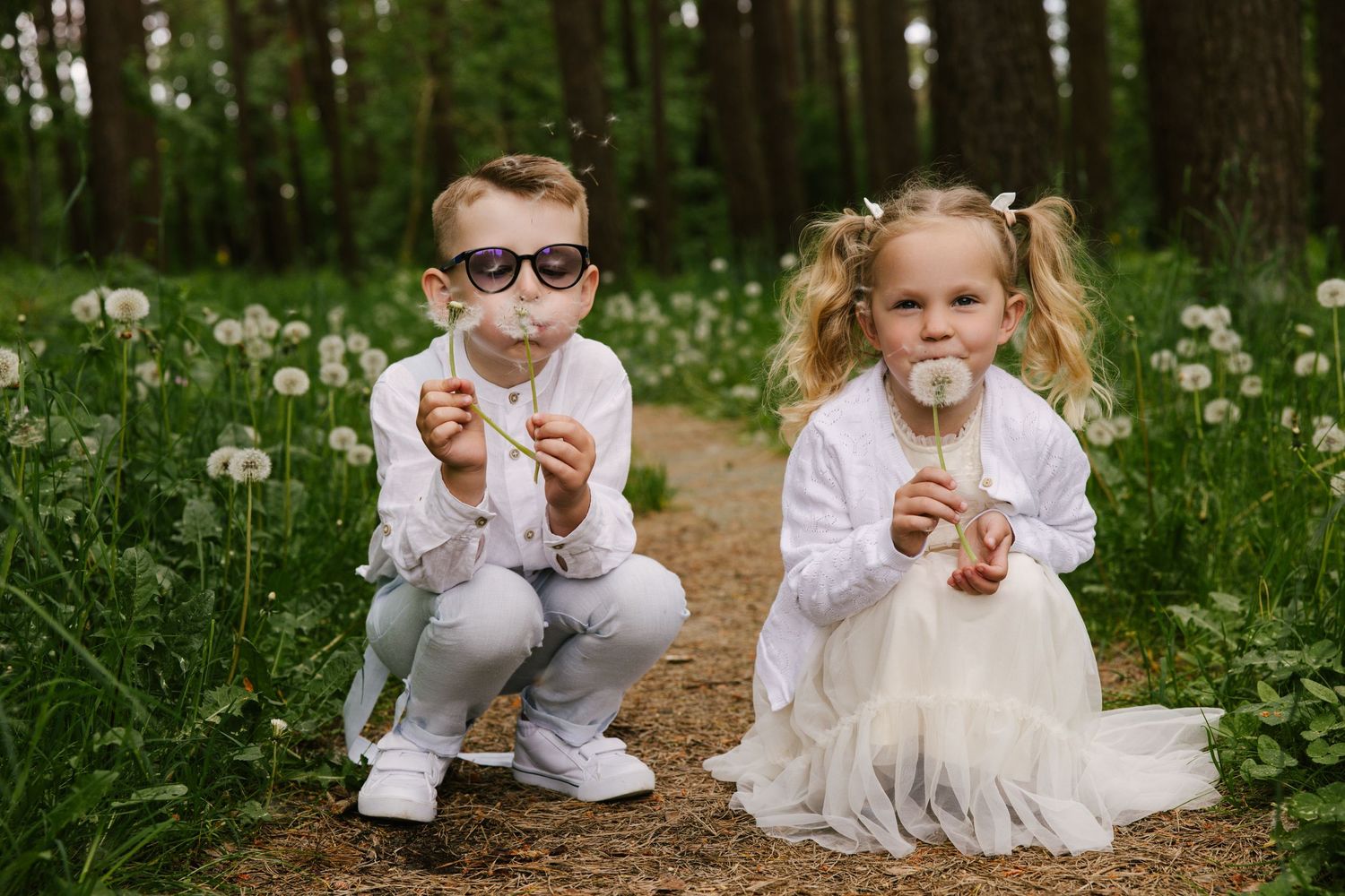 Children in white formal wear playing on a woodland trail among wildflowers.