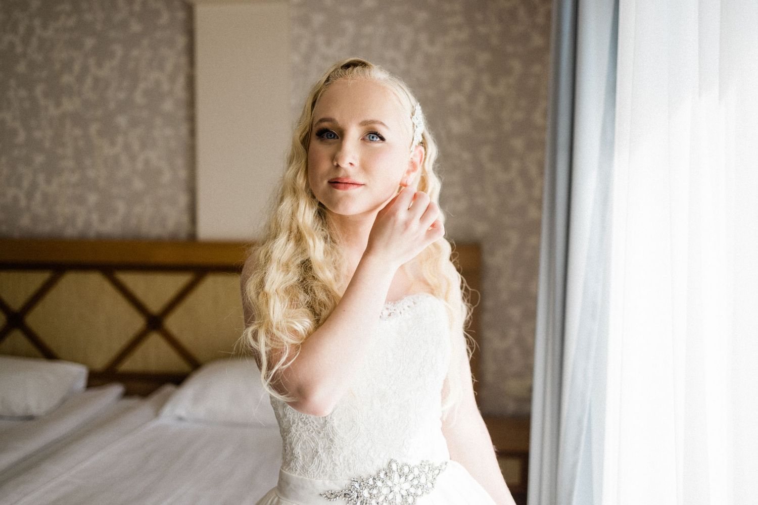 A bride in a lace wedding dress and veil poses by a window in a soft, natural light setting.