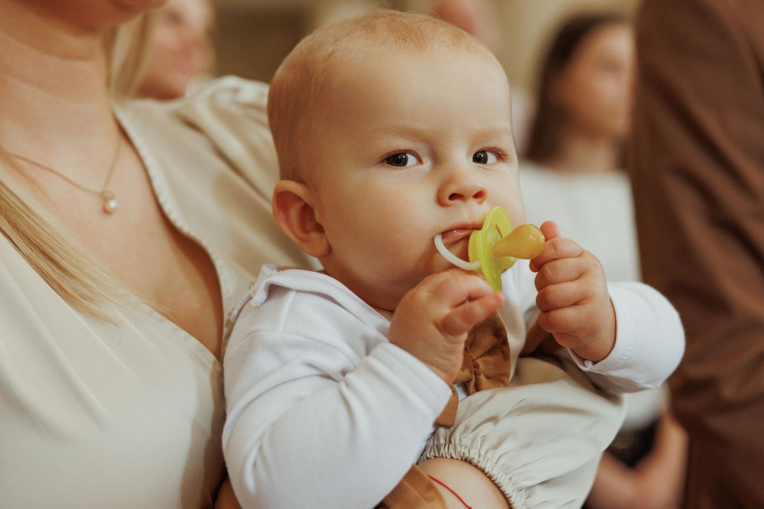 A baby in white clothing holds and tastes a piece of fruit while being held.