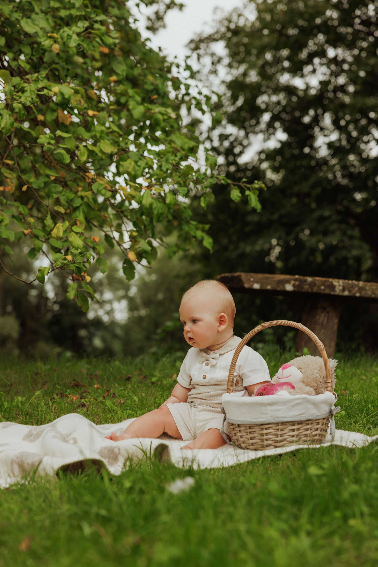 Baby in white outfit relaxes on blanket with wicker basket in grassy garden setting.