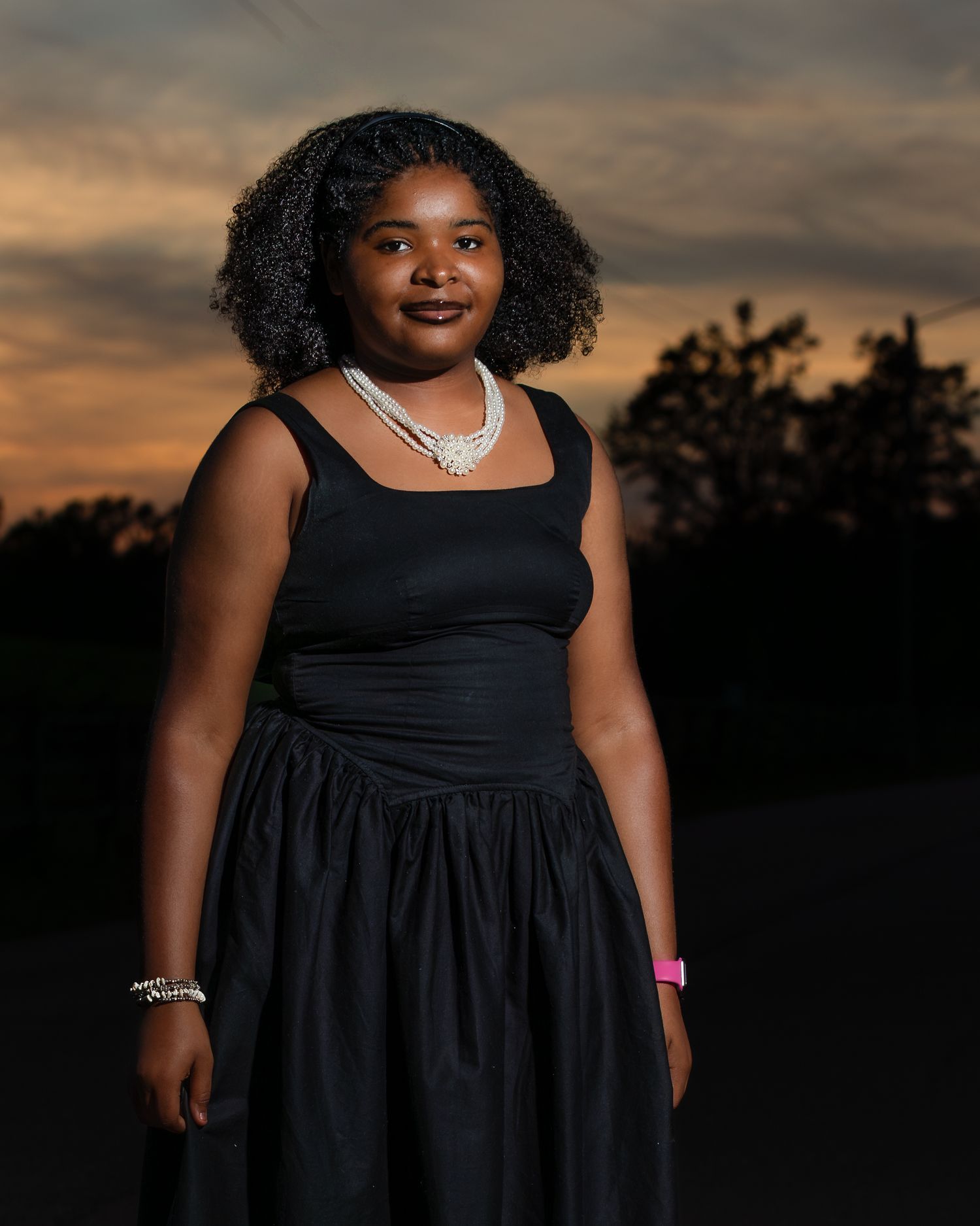 Young woman in black dress and pearl necklace poses outdoors at sunset with dramatic sky backdrop.