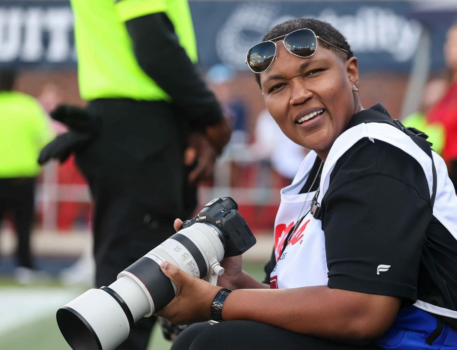A smiling woman holds a professional camera with a long telephoto lens at a sports event.