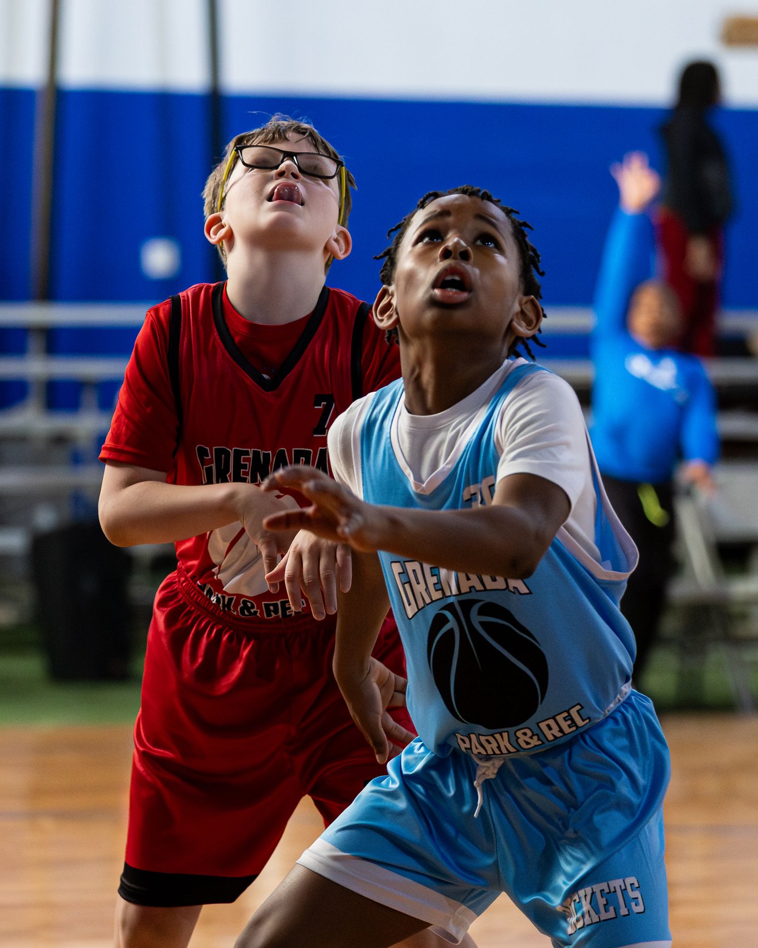 Two youth basketball players from opposing teams in red and blue jerseys compete for position under the hoop.