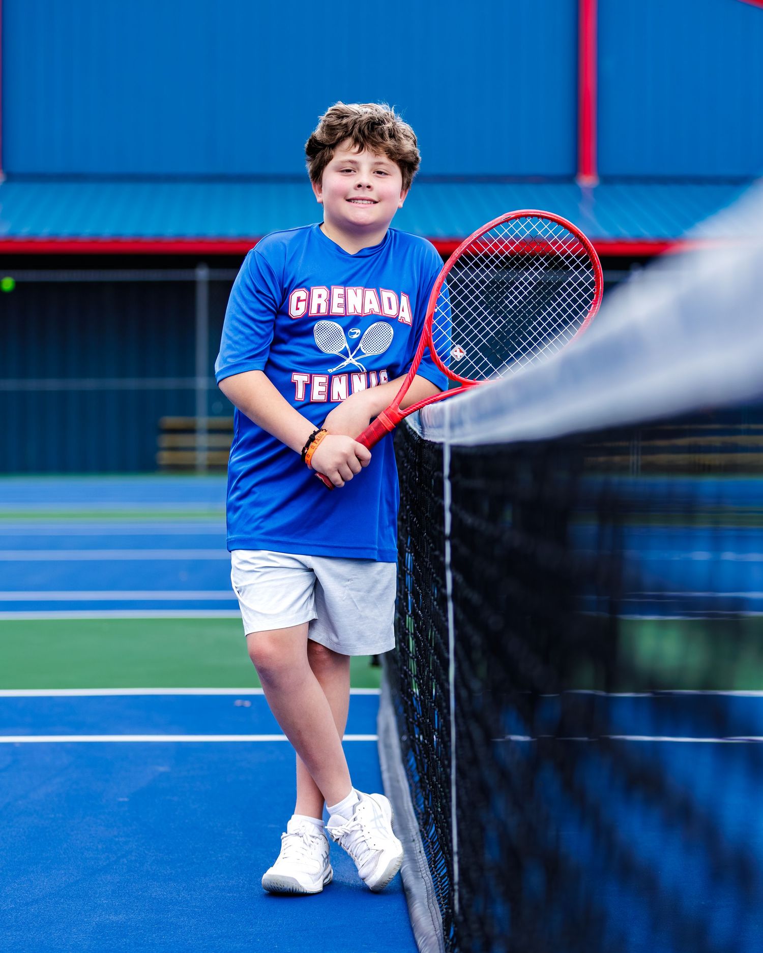 Young tennis player in a blue Grenada Tennis shirt stands next to the net on a bright blue court holding a red racquet.