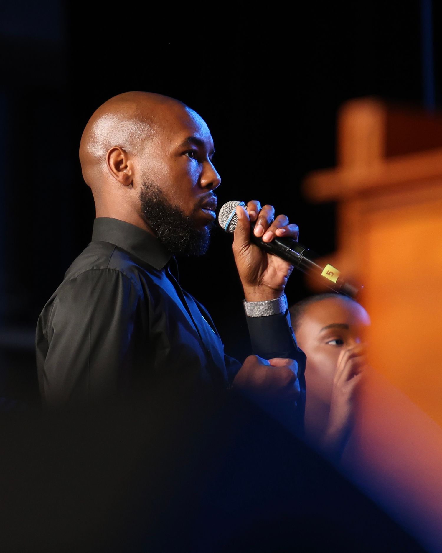 Performer singing on stage in dramatic lighting with silhouetted profile against dark background.