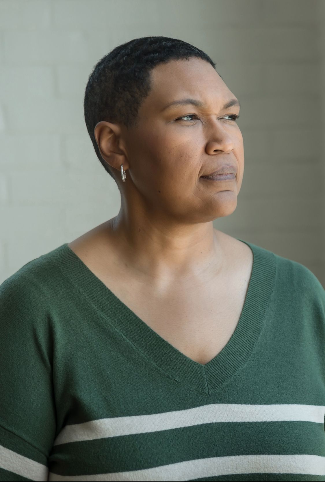 A person with short hair wearing a green v-neck sweater gazes thoughtfully to the side against a light background.