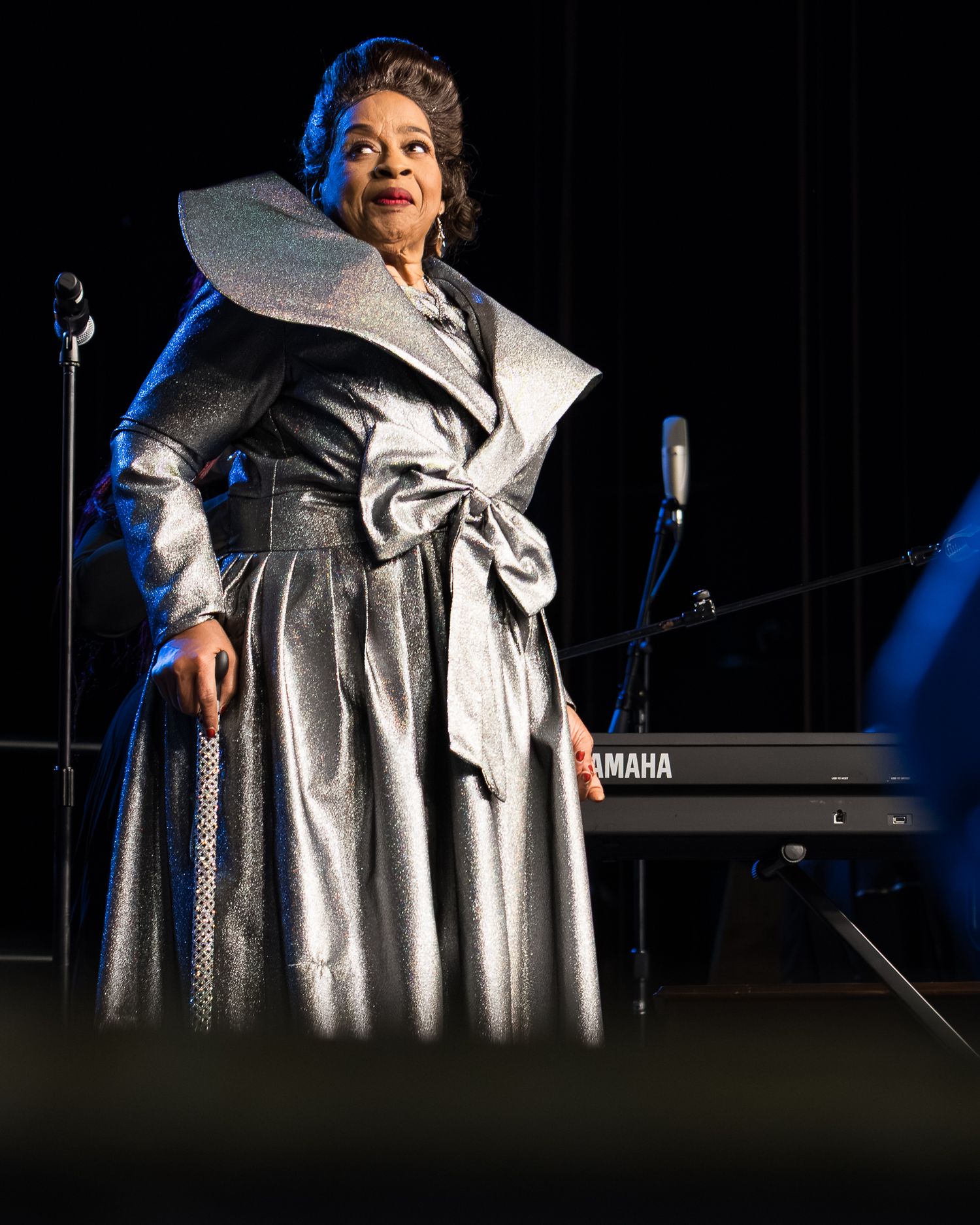 A performer in a sparkly silver dress with a dramatic bow detail stands on a dimly lit stage with blue lighting.