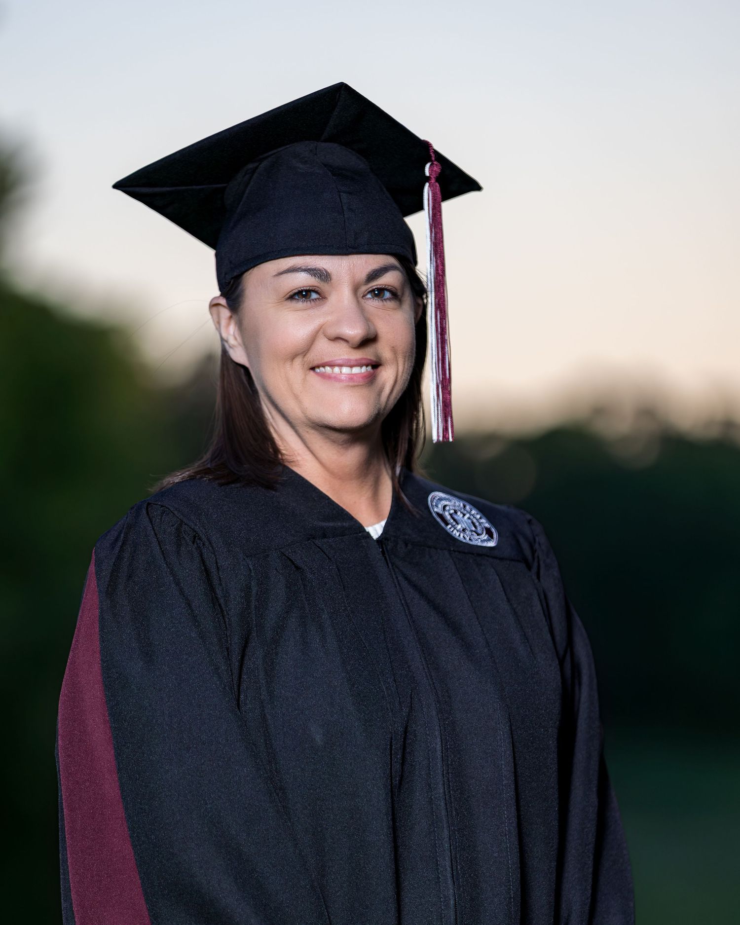 Woman in black graduation cap and gown smiling outdoors at dusk during commencement ceremony.