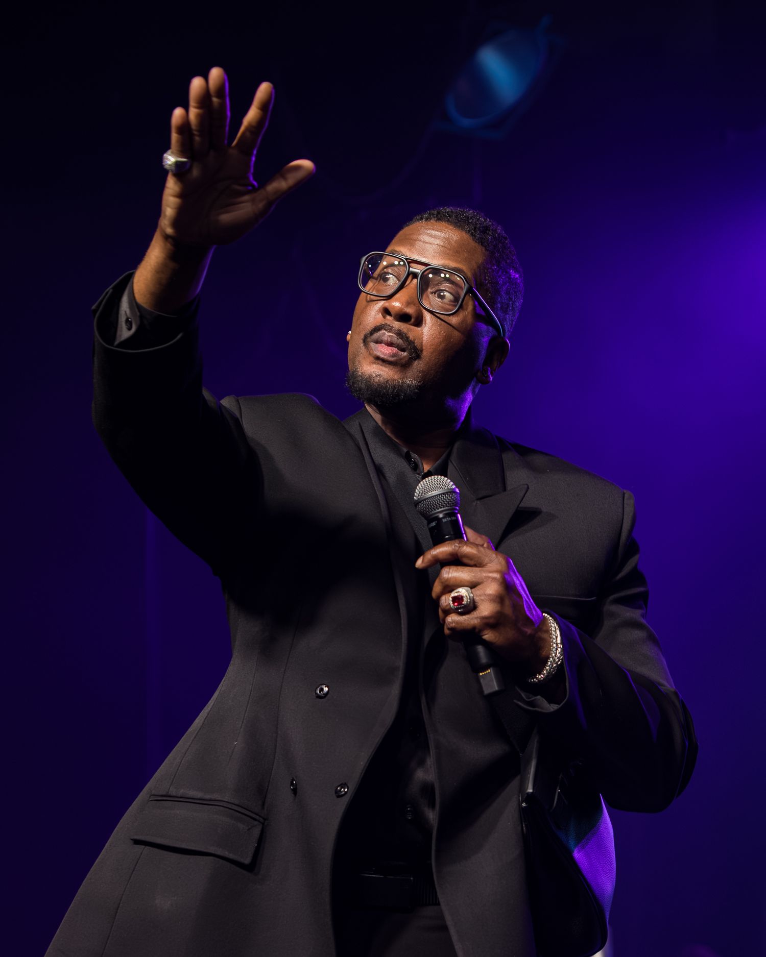 Man in black suit holding microphone gestures on stage with purple lighting during live performance.