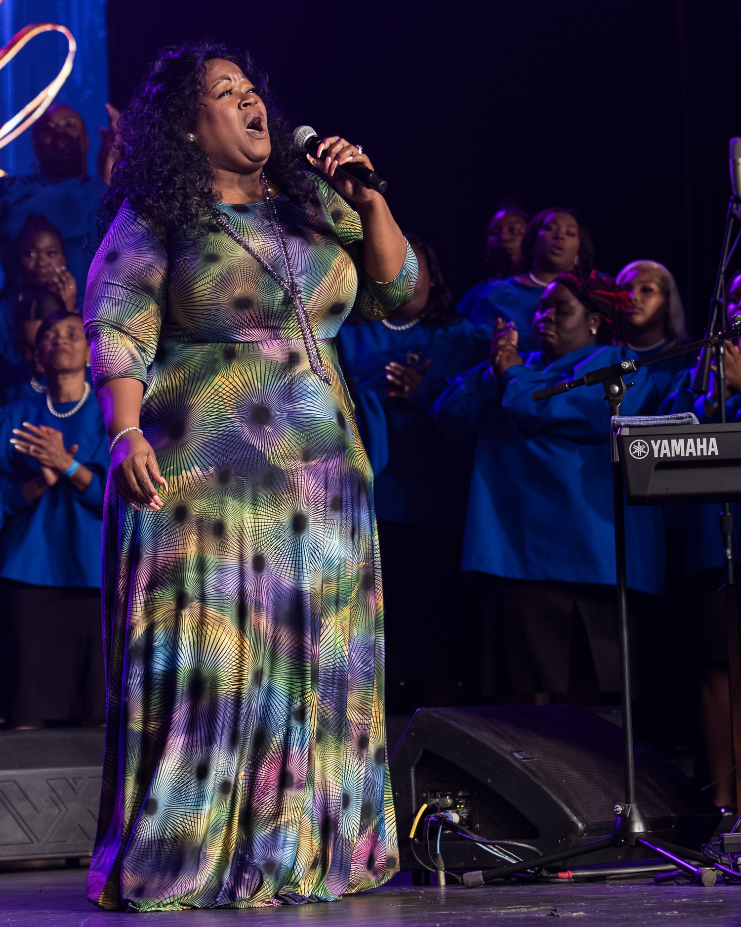 A performer in a colorful tie-dye dress sings passionately on stage with a microphone during a concert performance.