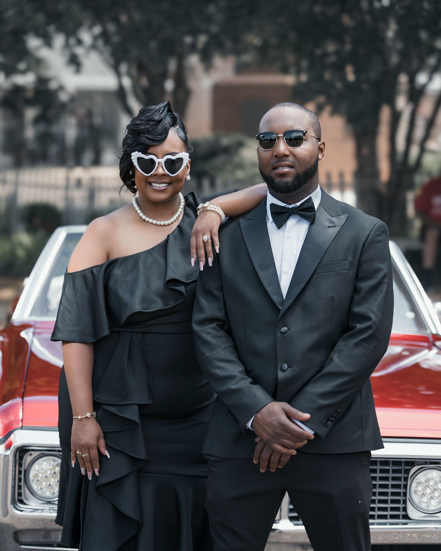 Stylish couple in formal black attire pose together against a vintage red car for elegant photos.