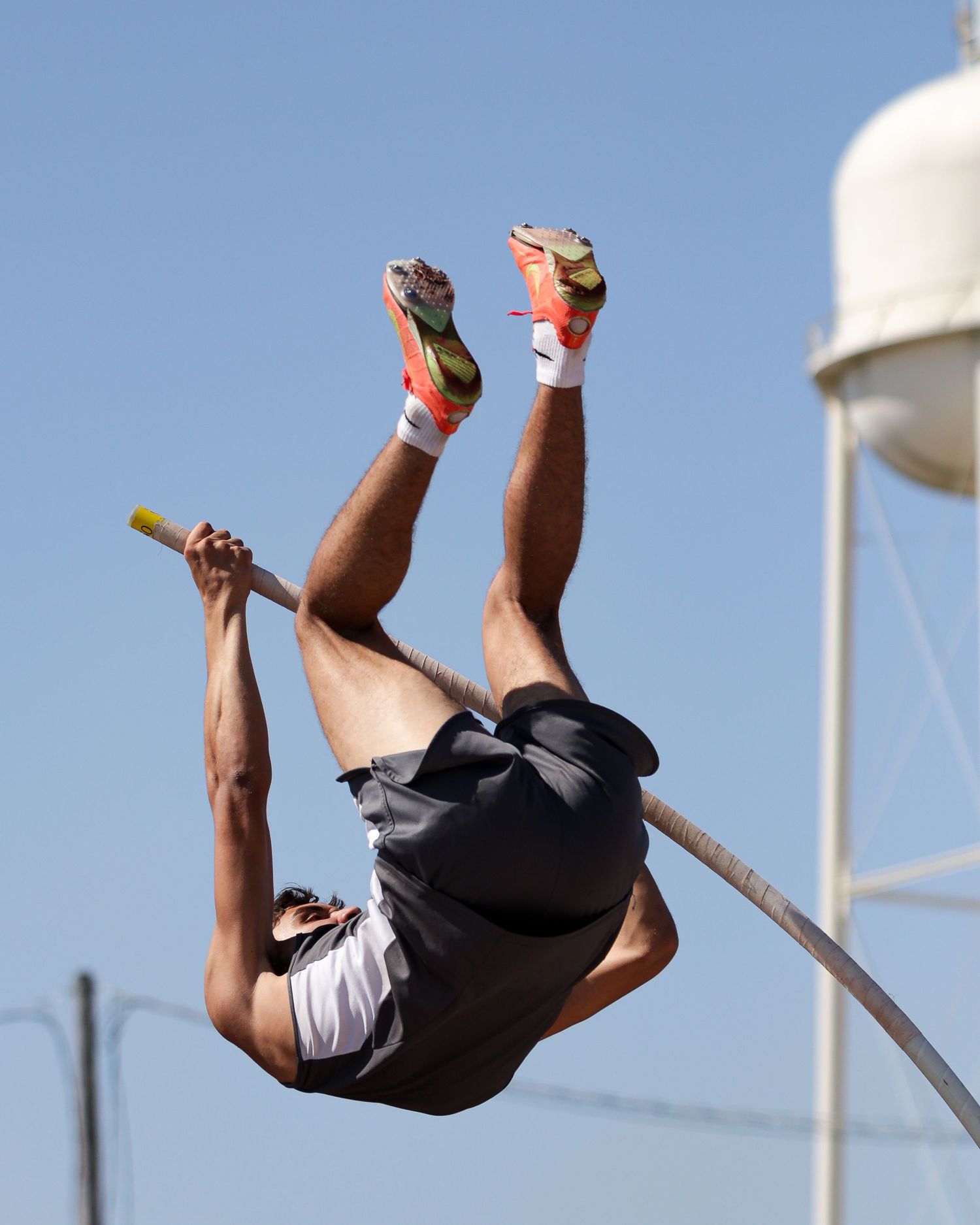 Athlete performing pole vault against blue sky with water tower in background.