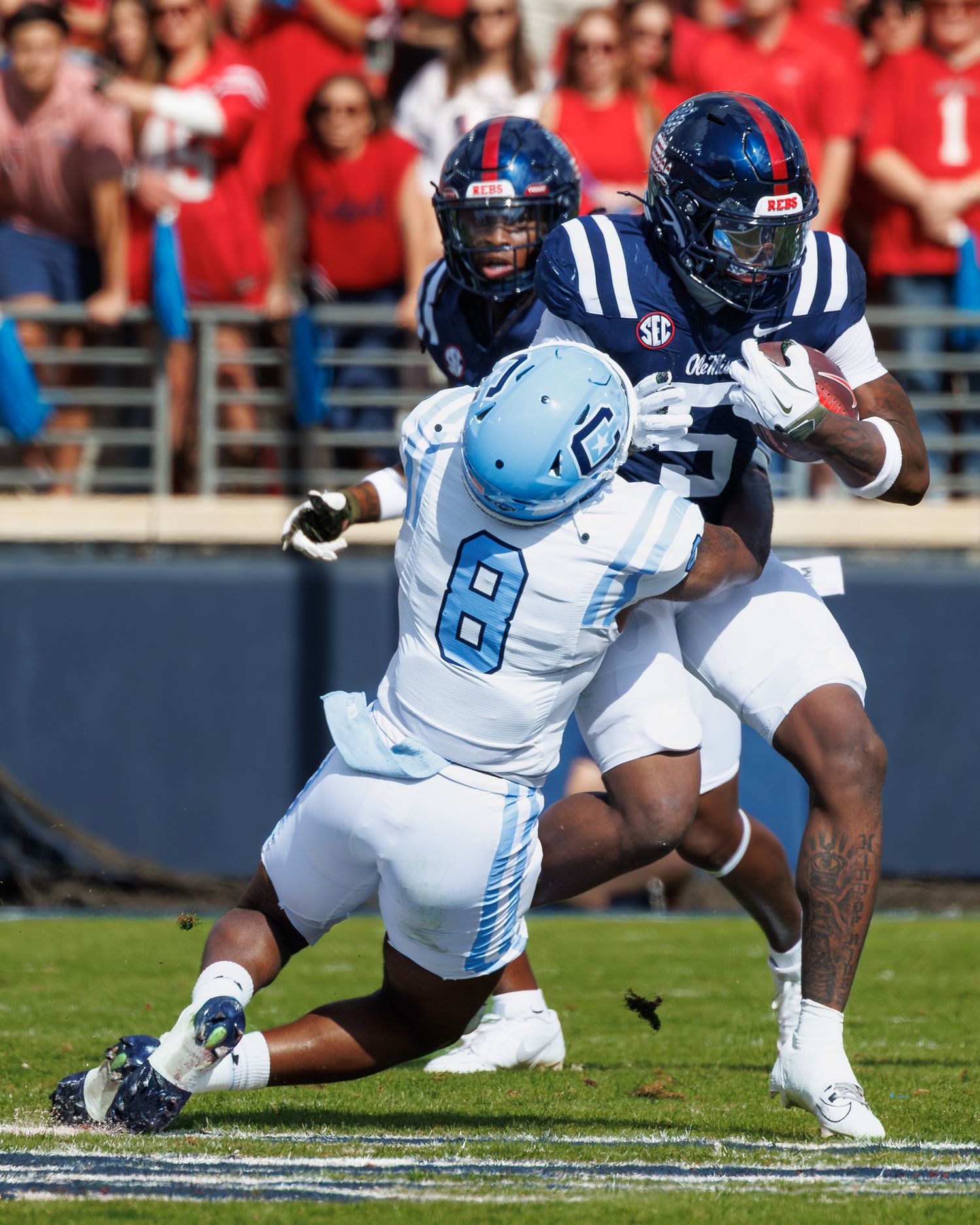 A football player in light blue jersey falls forward during a tackle in a game against navy blue uniformed opponents.