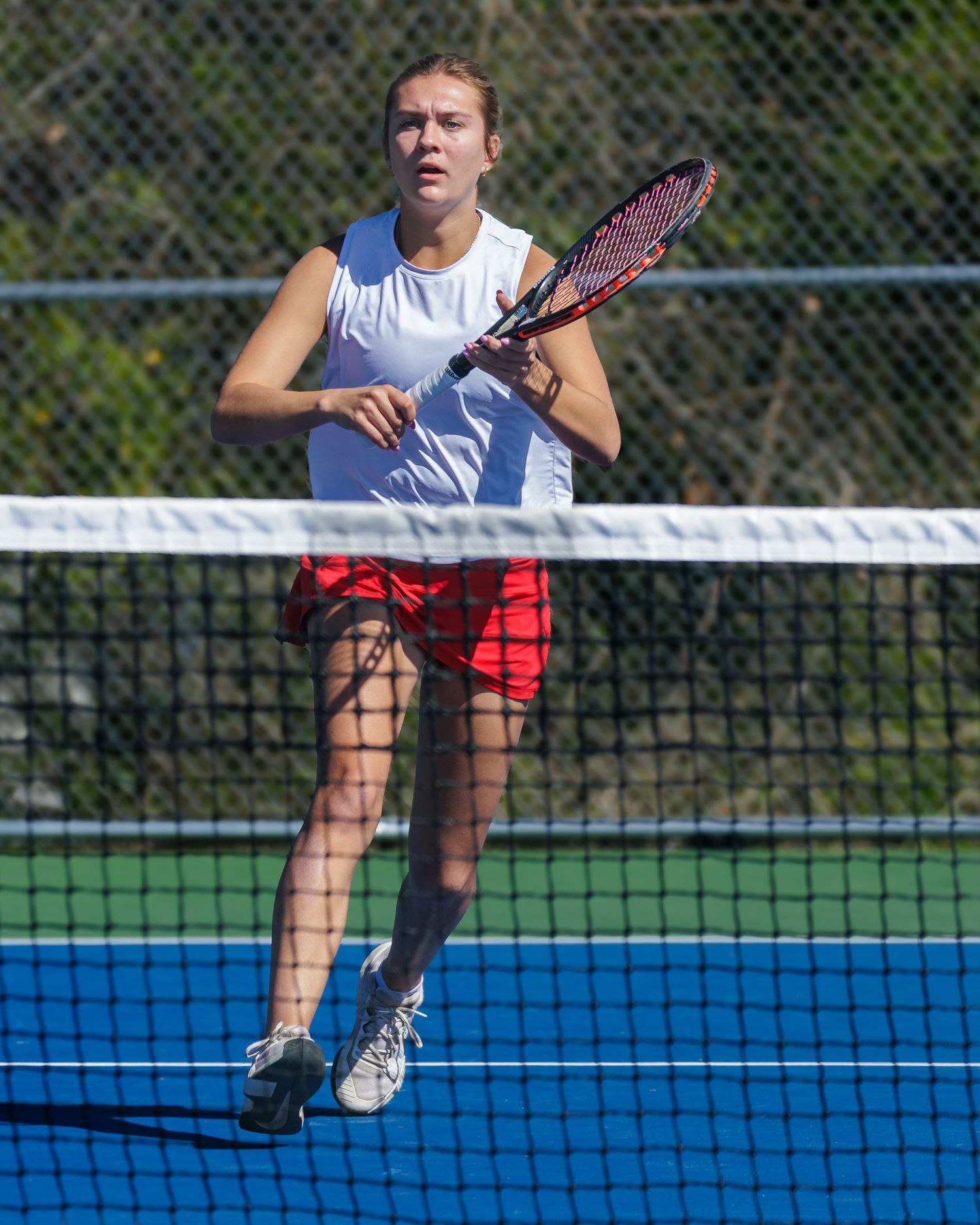 Tennis player in white tank top and red shorts stands at net during match on outdoor blue court.