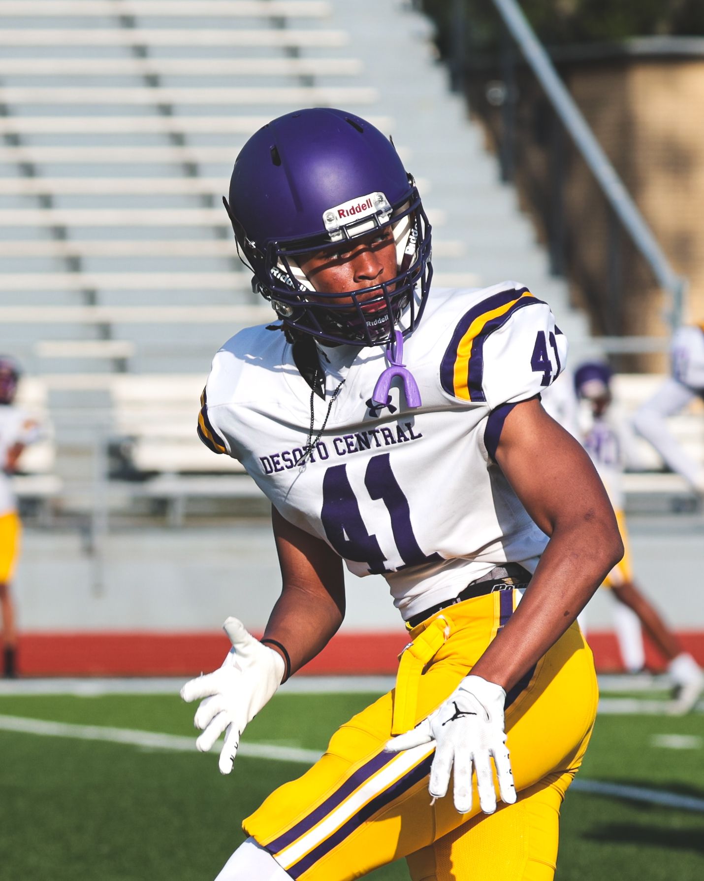 Players in purple and yellow uniforms practice defensive moves on the football field.