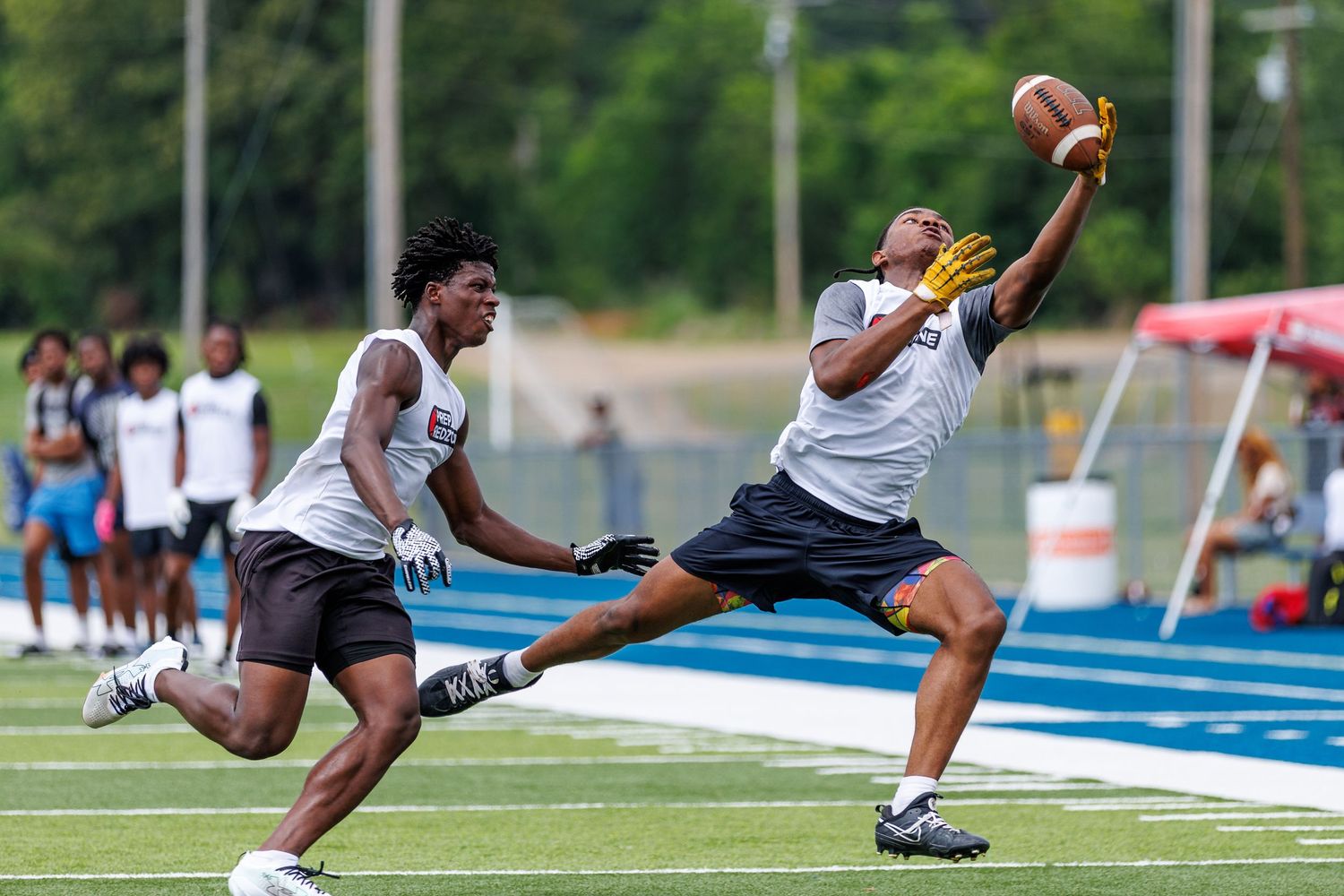 Two football players compete for a catch during an outdoor game on a blue turf field on a sunny day.