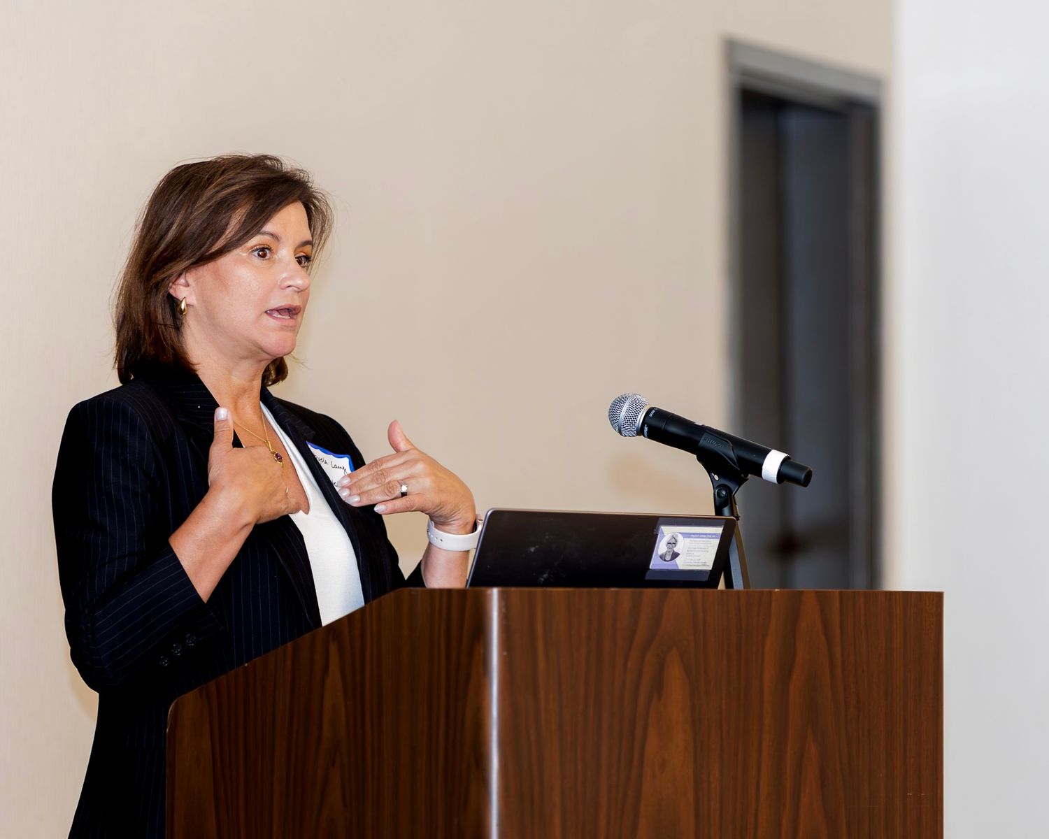 Professional woman in black blazer speaking at wooden podium with microphone during business conference presentation.