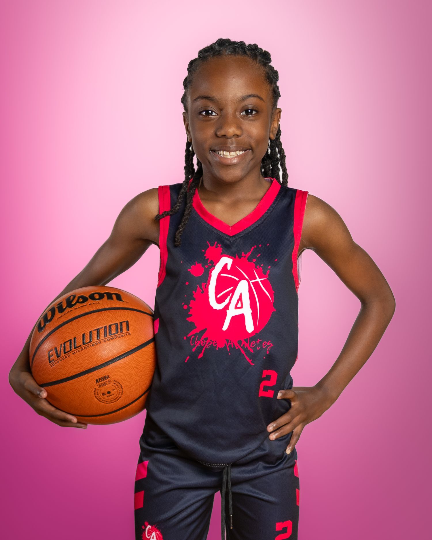Young girl in black and red CA basketball uniform holding a Wilson Evolution ball, smiling against pink background.