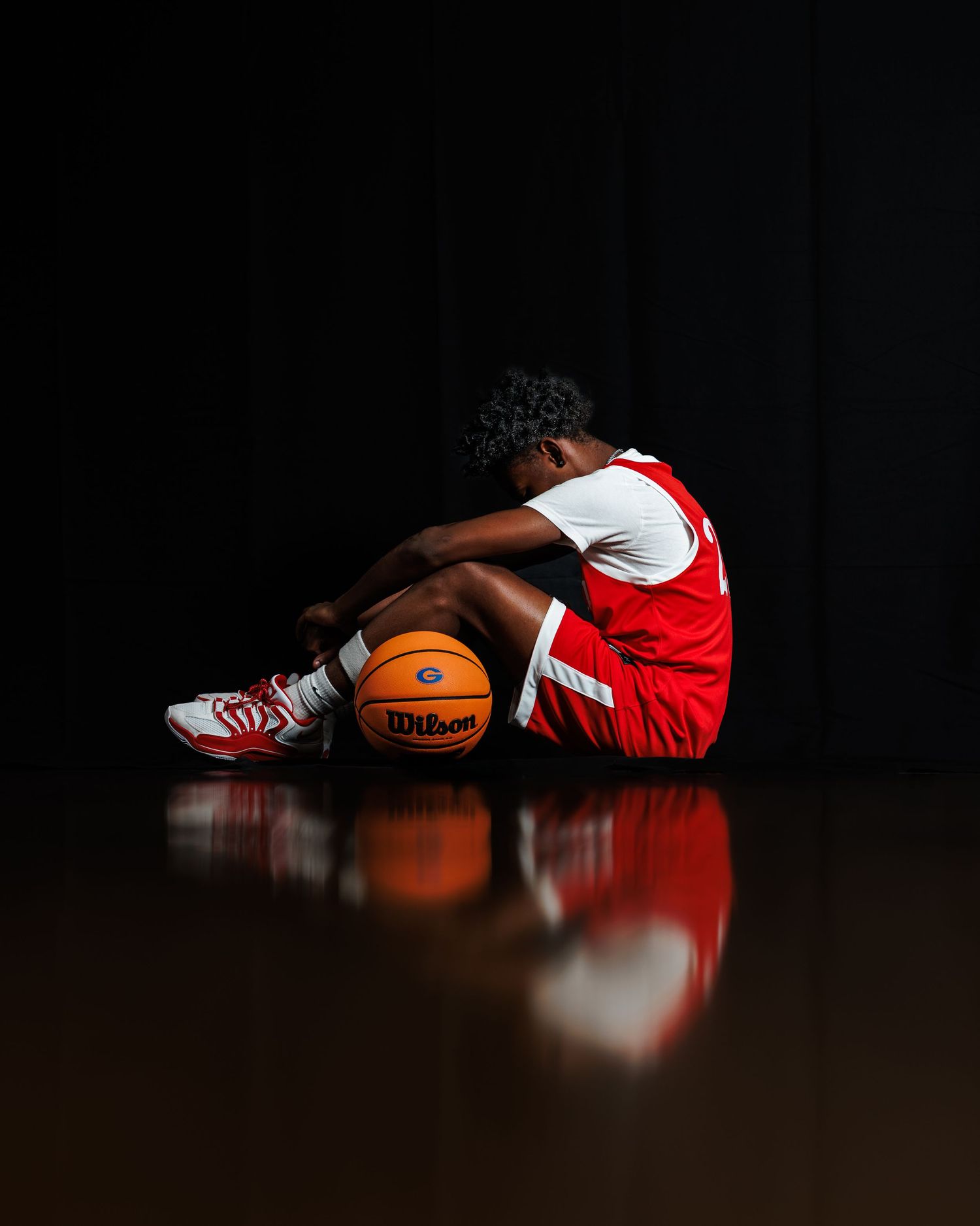 Basketball player in red uniform sitting on court with Wilson ball, head down, dramatic dark background.