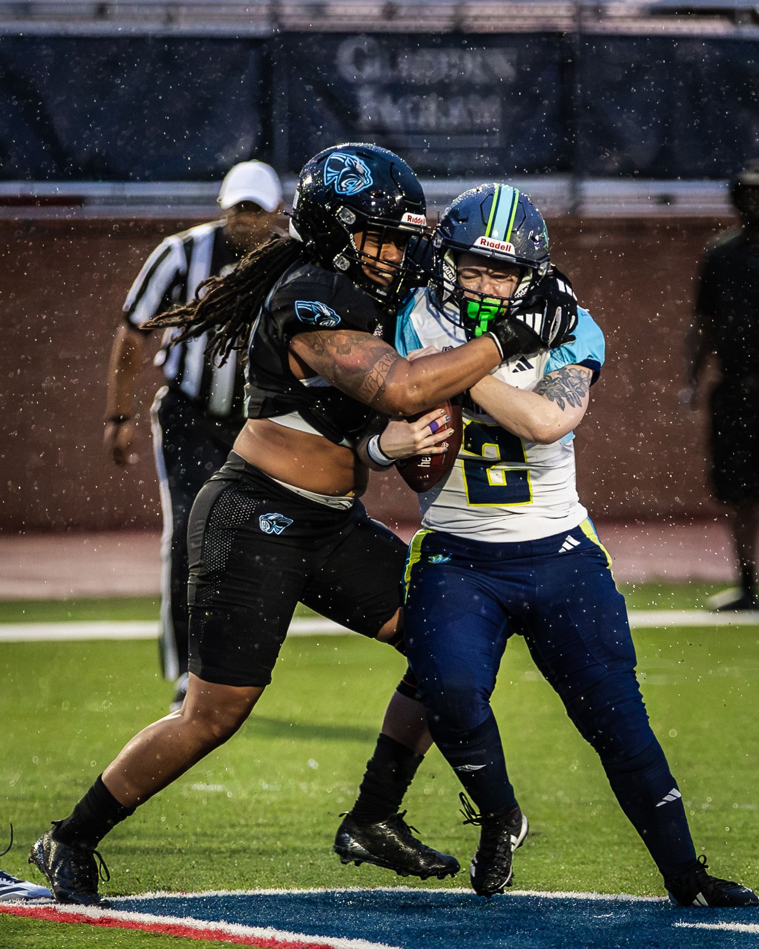 Two female football players compete in a rainy night game, one in black uniform tackling the other in teal and navy.