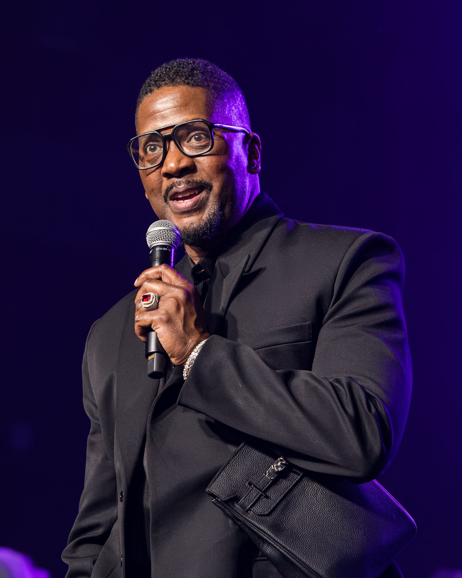 A performer in a black suit and glasses holds a microphone while singing on stage with purple lighting in the background.