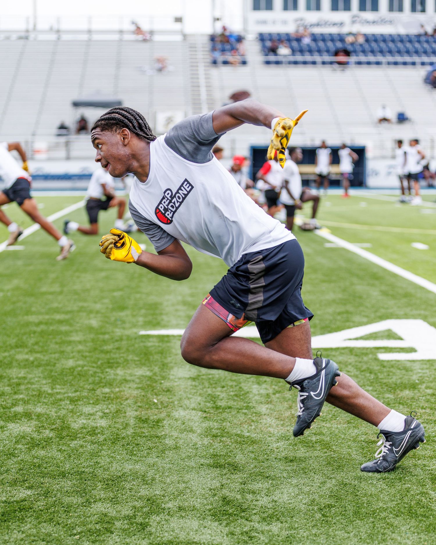 Athletic football player sprinting on a stadium field during a training event, wearing gloves and cleats.