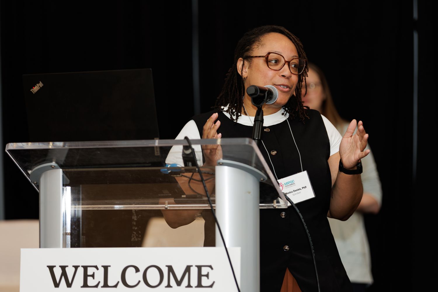 Woman with glasses speaking at a podium with a Welcome sign at a professional conference event.