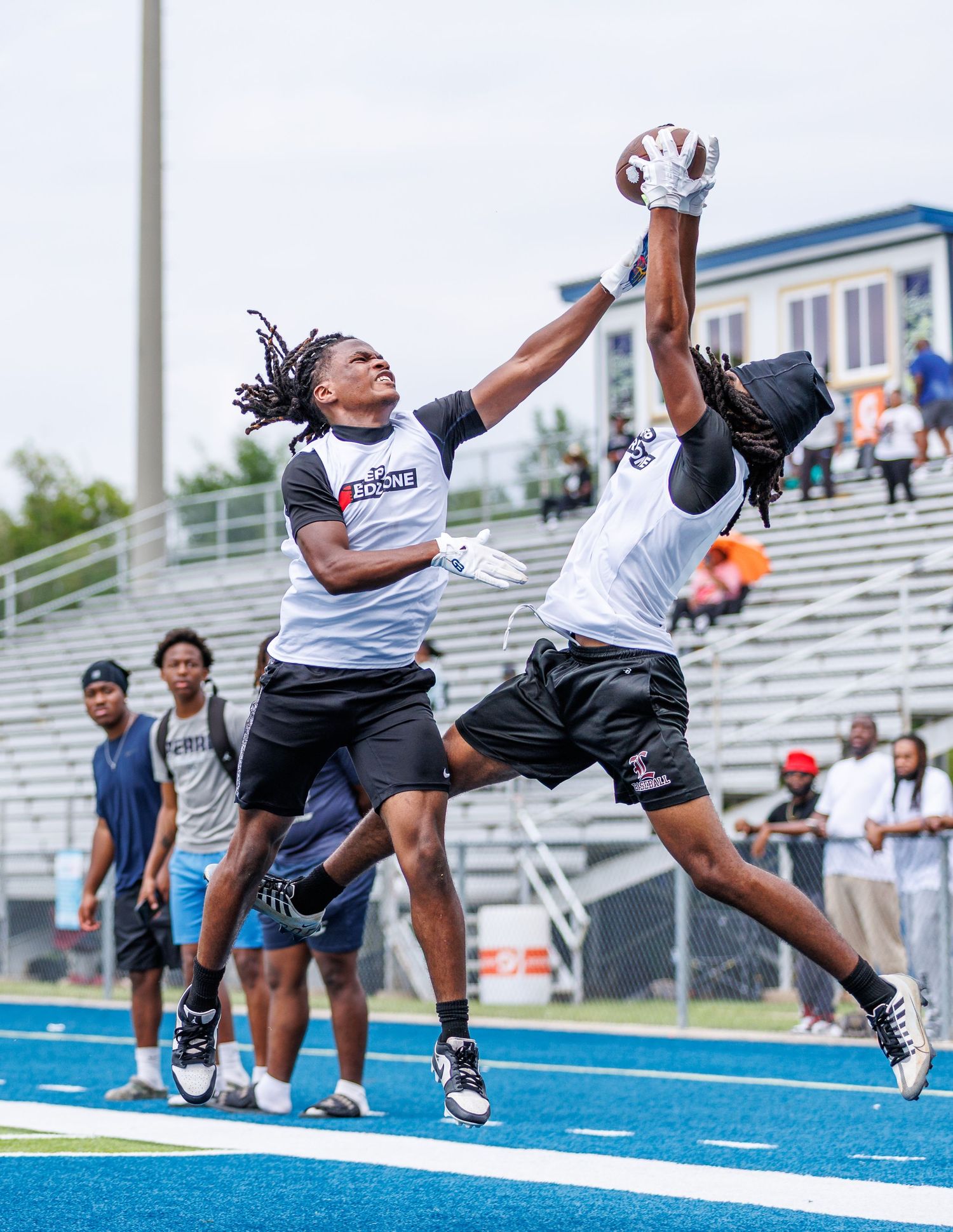 Two football players jump to contest a pass during an outdoor 7-on-7 tournament on a blue turf field.
