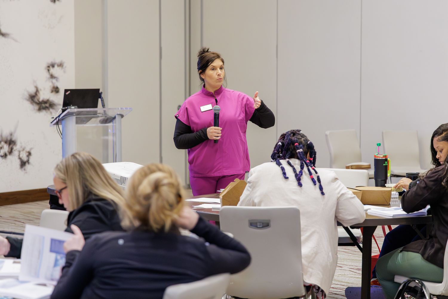 Healthcare professional in pink scrubs presenting to a group of seated attendees in a conference room training session.