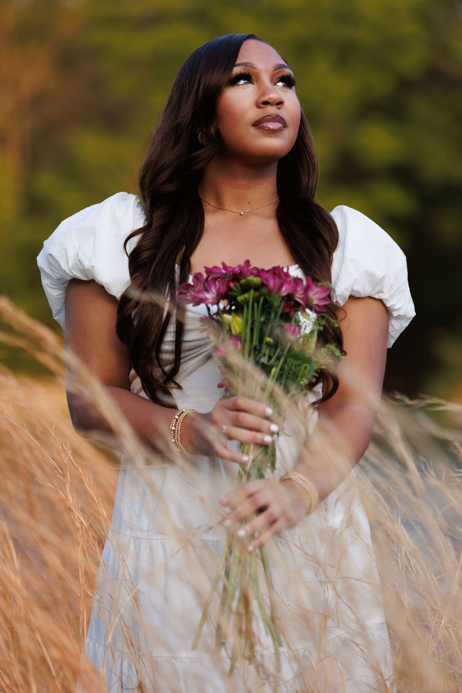 Woman in white dress holding pink flowers in a golden grass field, gazing upward with long dark hair.