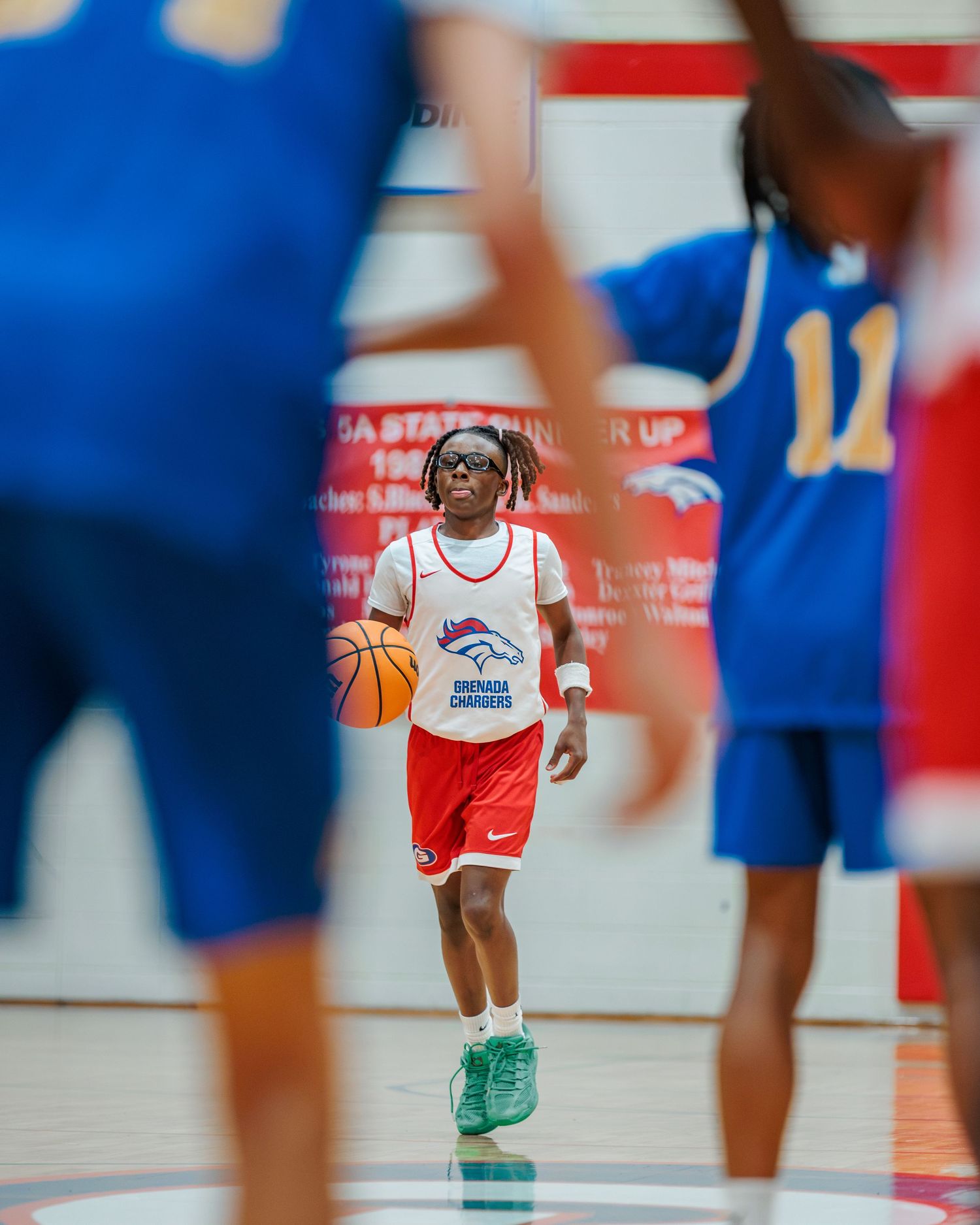 Young basketball player in red Denver Broncos jersey dribbles down court during indoor game.