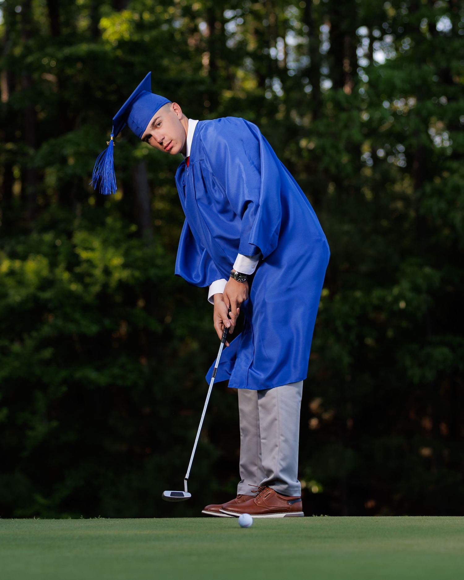 Graduate in blue cap and gown putting on golf green, surrounded by lush trees in natural light.