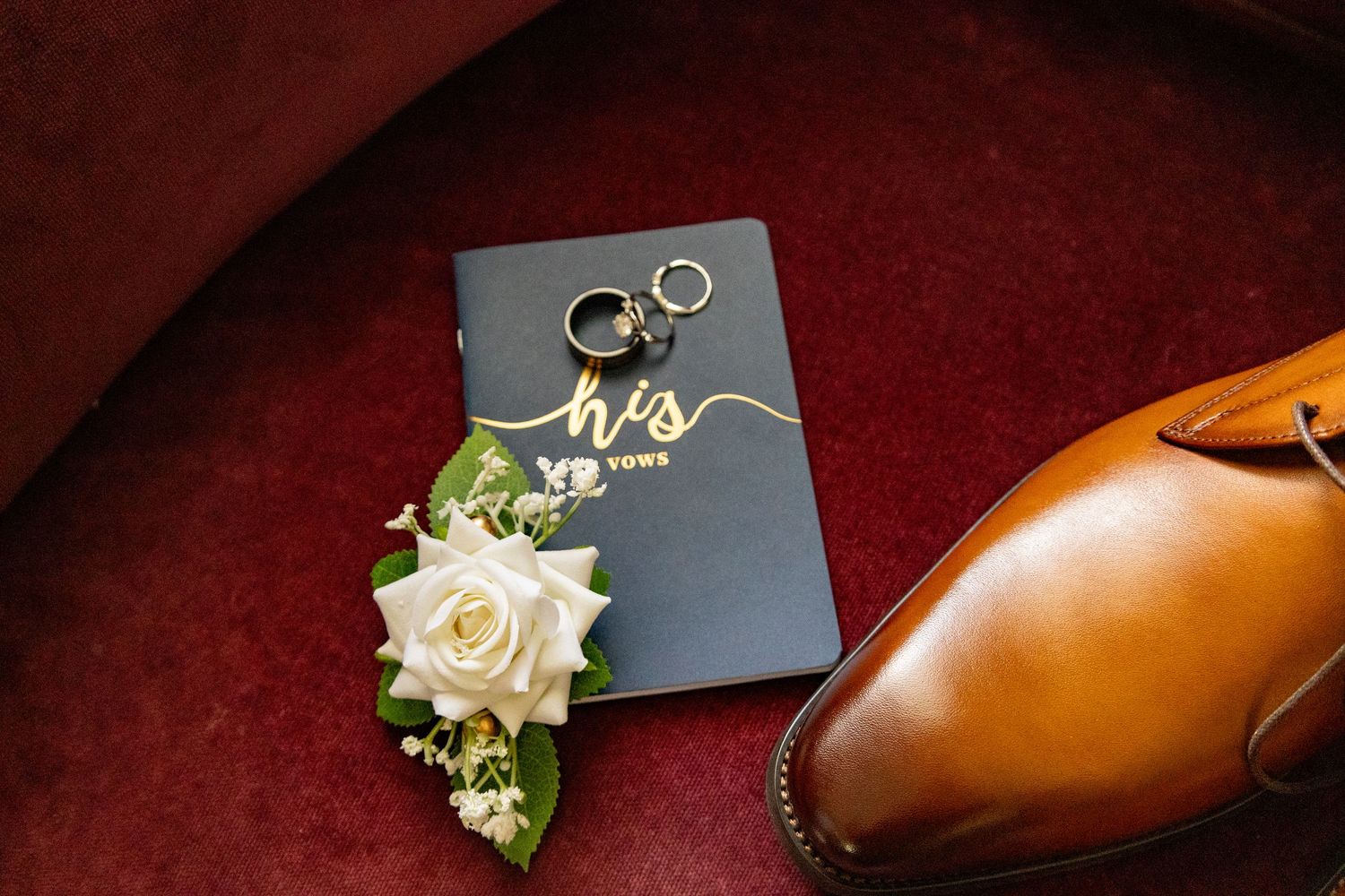 Wedding vows book with white rose and brown leather shoe on burgundy surface.