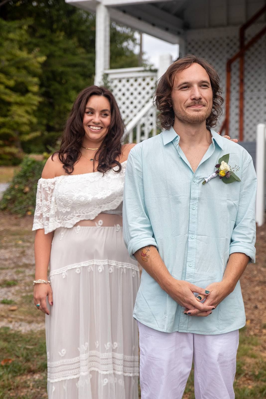 A couple in casual wedding attire stands together in front of a white latticed porch.
