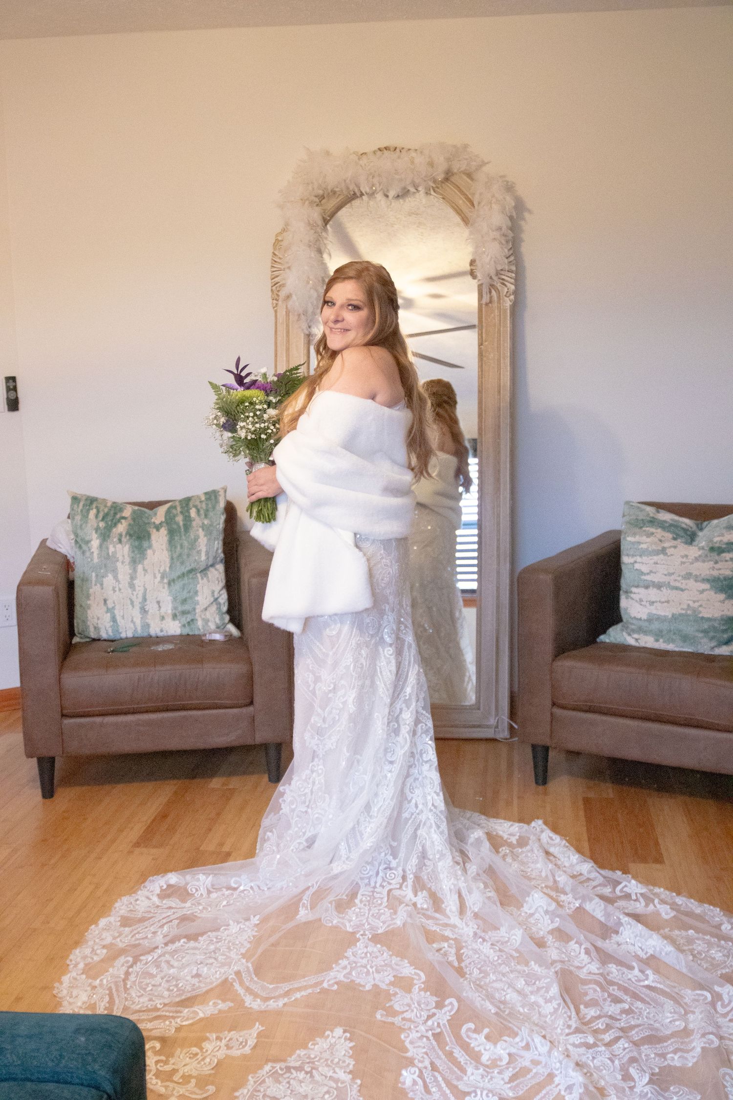 An elegant white lace wedding gown with a long train is showcased in a room with brown armchairs and teal throw pillows.