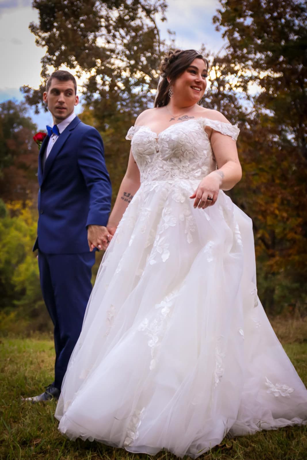 A wedding couple poses outdoors, with the bride in a lace ballgown and groom in navy suit against autumn foliage.
