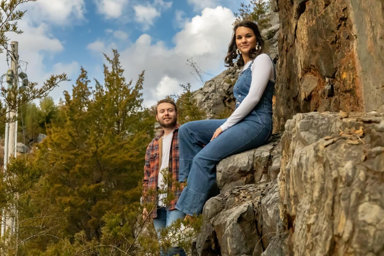Adventure couple posing on rocky cliff edge with evergreen forest and cloudy sky backdrop.
