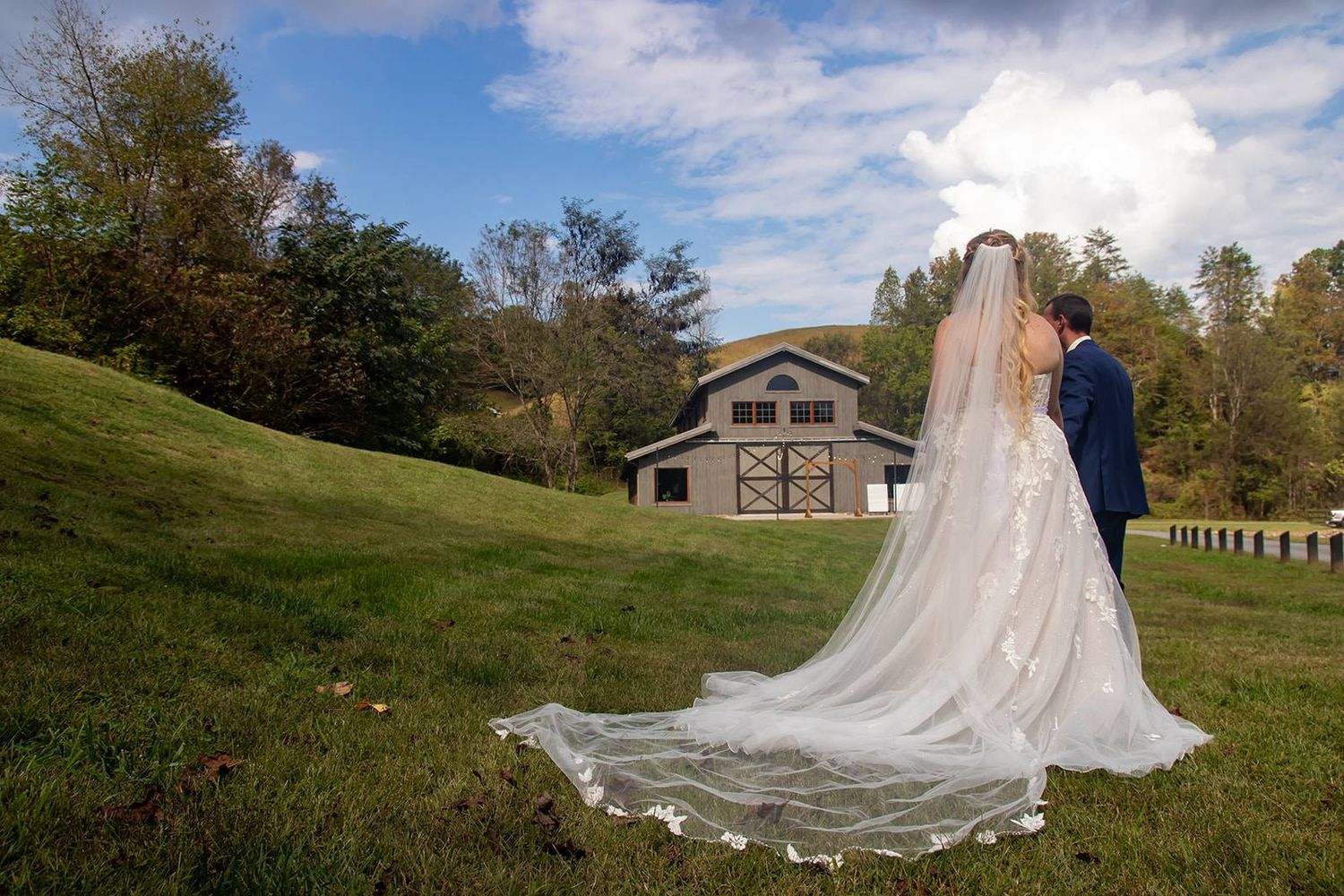 A bride in a flowing veil stands with someone on a grassy hill with a rustic building in background.