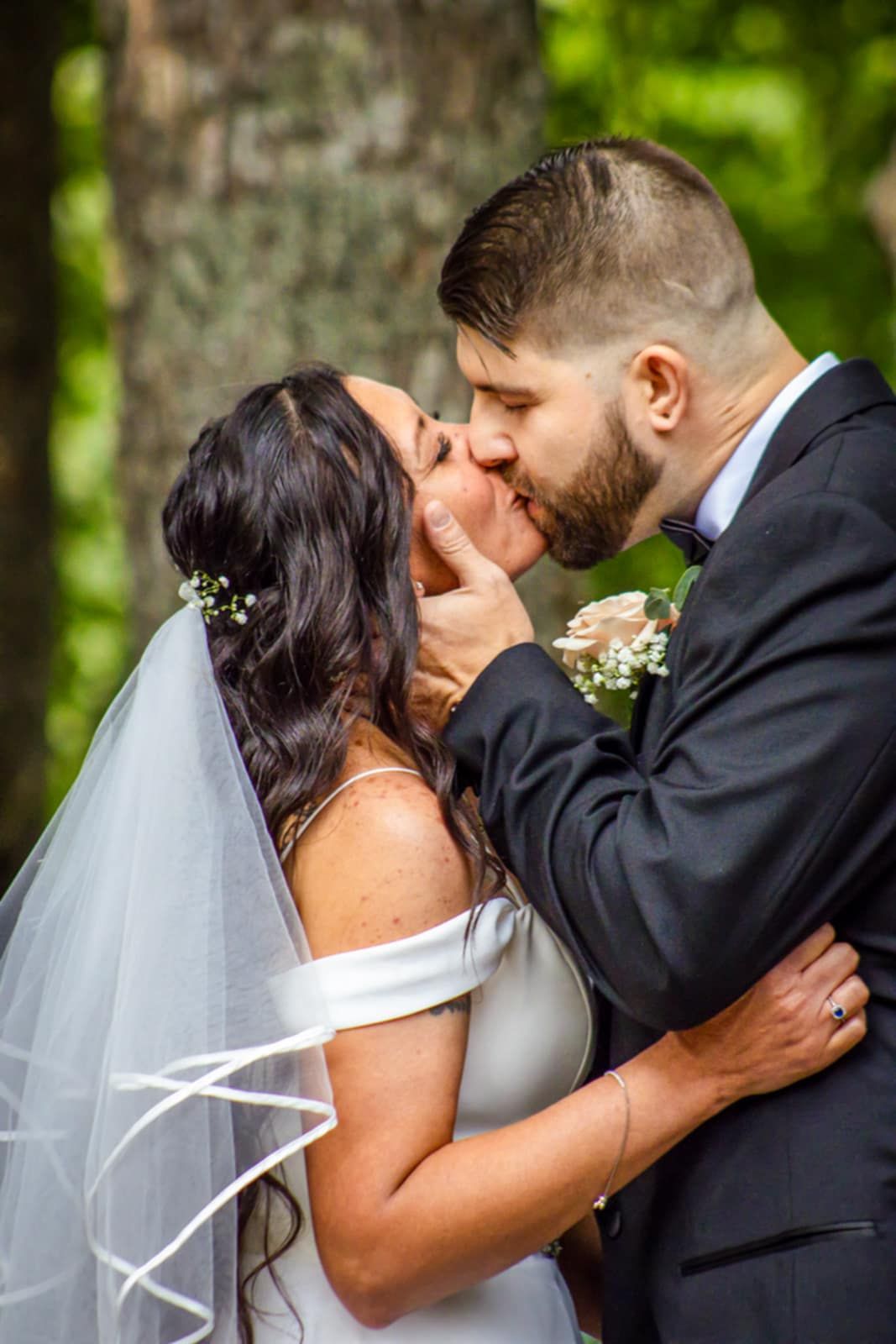 A tender wedding kiss moment captured in a natural woodland setting.