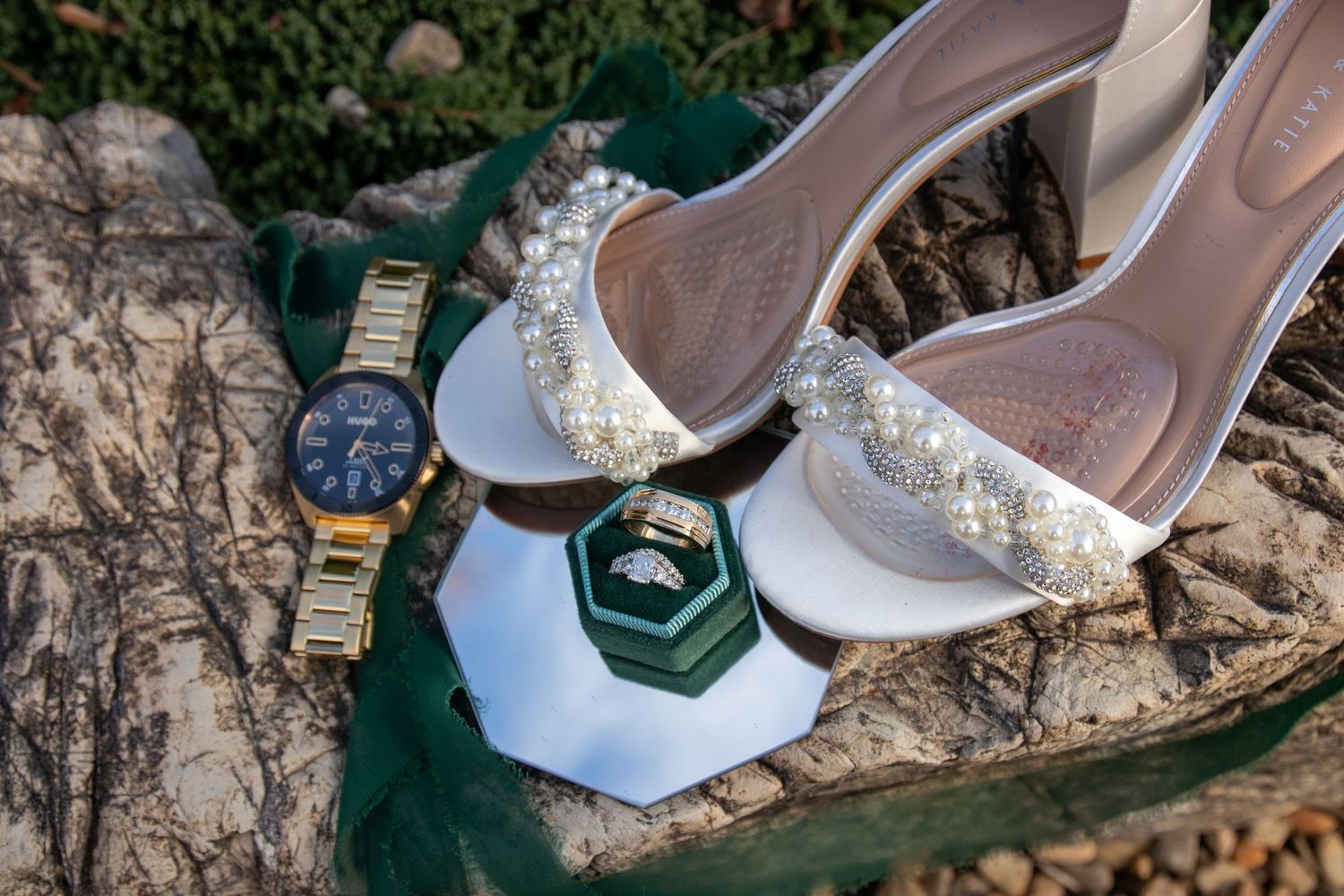Wedding accessories including pearl-adorned heels, rings, and watch arranged on natural stone.