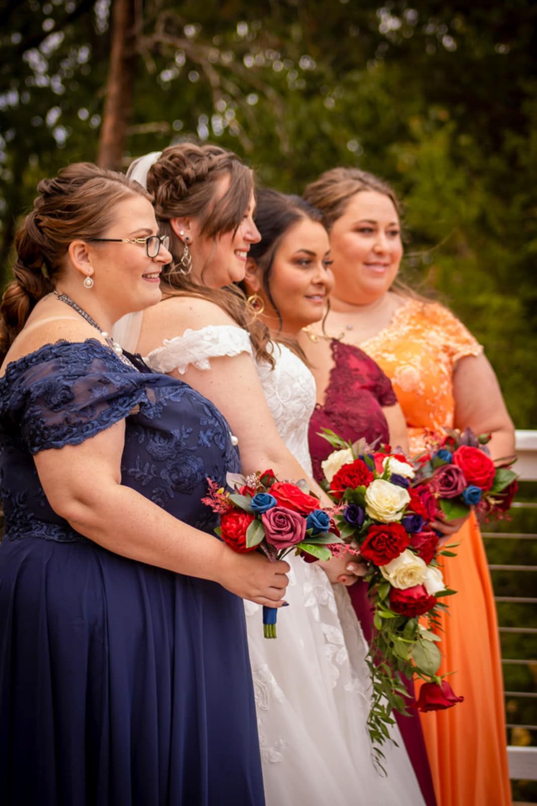 Bridesmaids in navy and orange dresses hold red and white bouquets while standing with the bride.