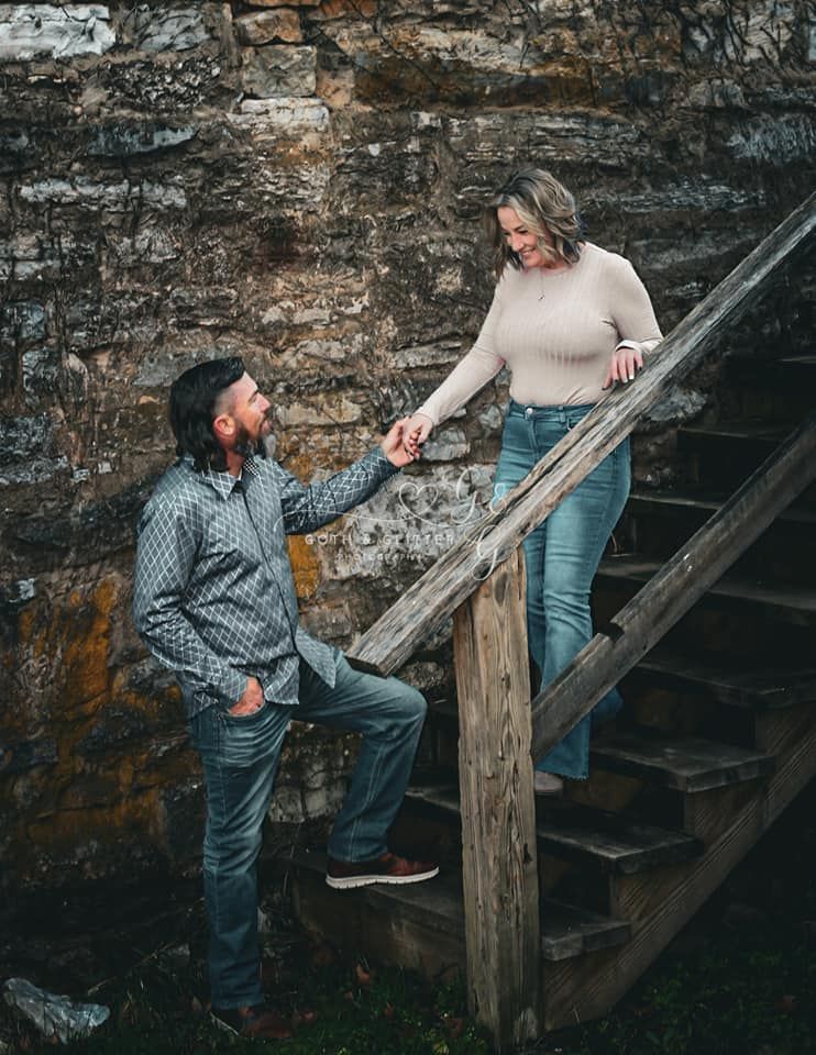 A couple holds hands on rustic wooden stairs against an old stone wall.