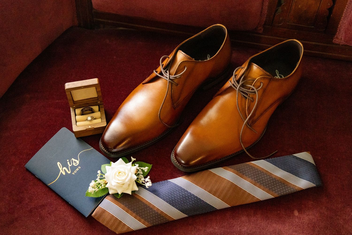 Brown leather dress shoes with wedding rings and striped tie on burgundy surface.
