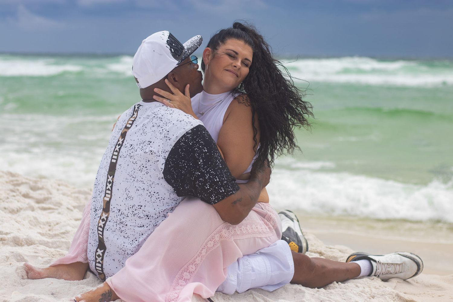 Romantic couple embracing on sandy beach with turquoise ocean waves in background.