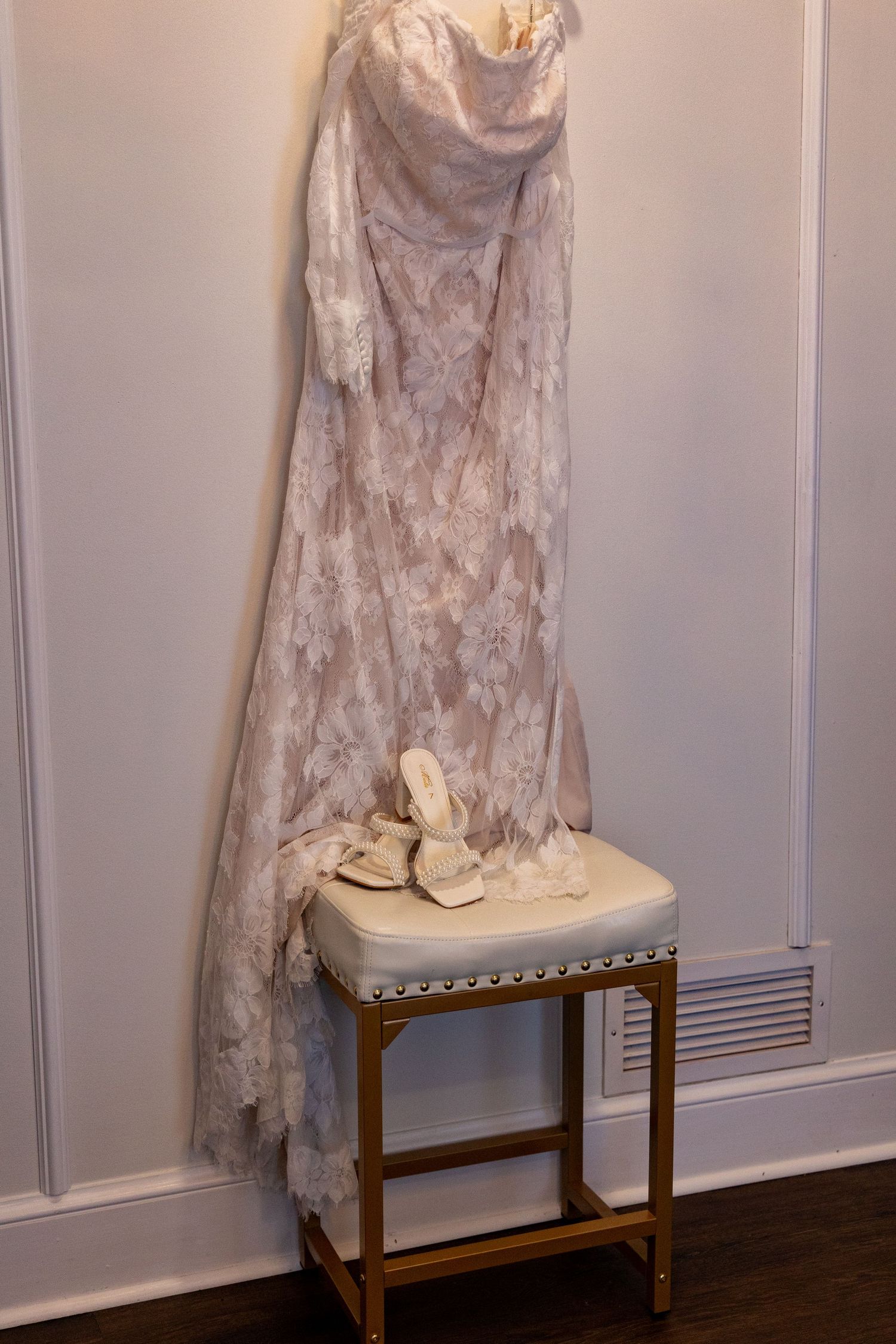 Vintage lace wedding dress hanging above small upholstered stool against white wall.