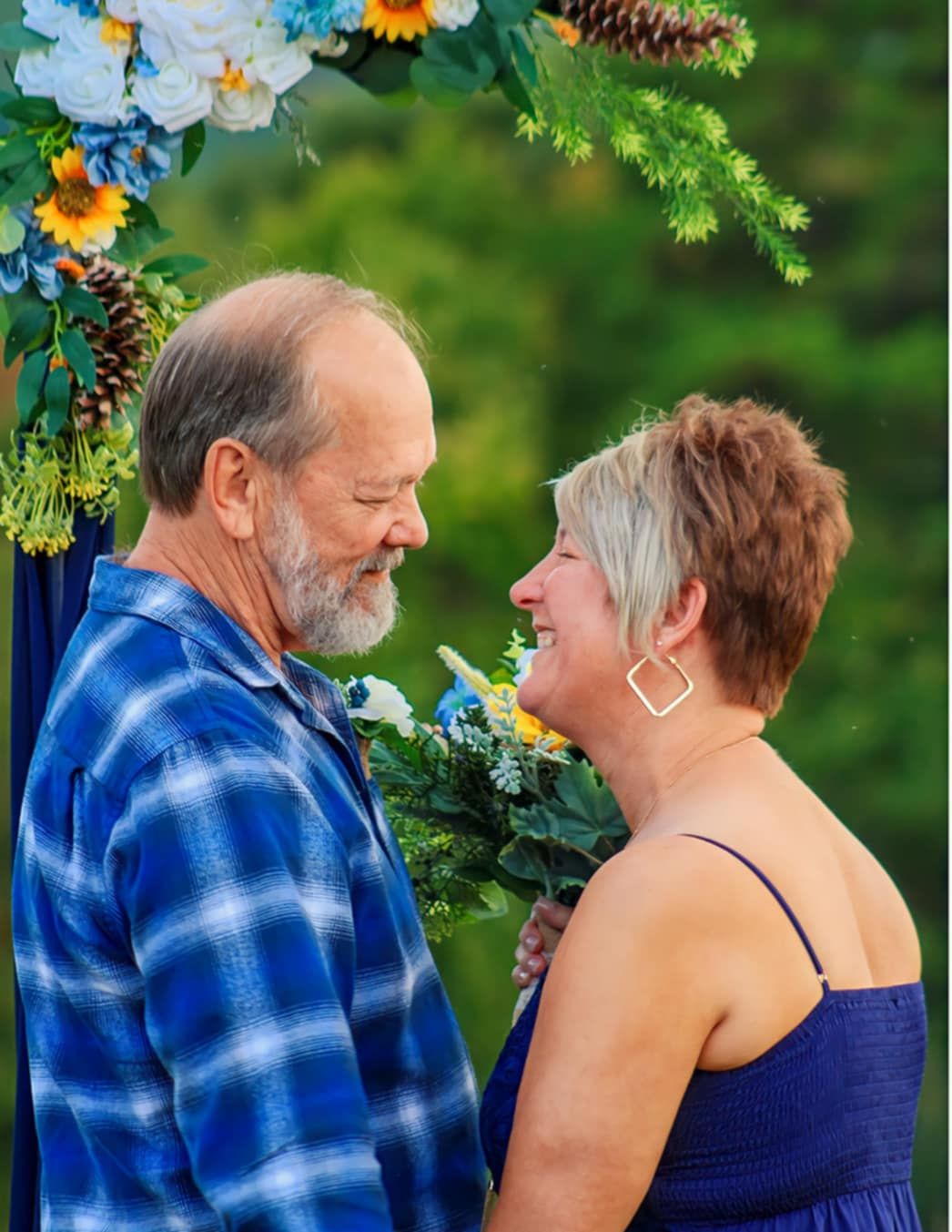 A couple stands beneath a floral arch with sunflowers and pinecones during an outdoor ceremony.